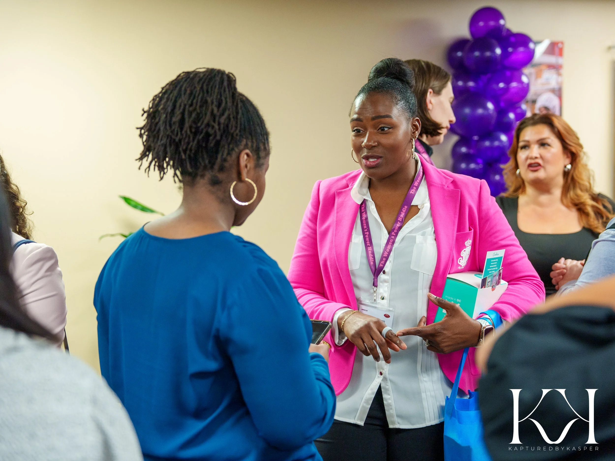 Two women engaged in conversation at a social event, one in a pink blazer and the other in a blue top, with purple balloons in the background.