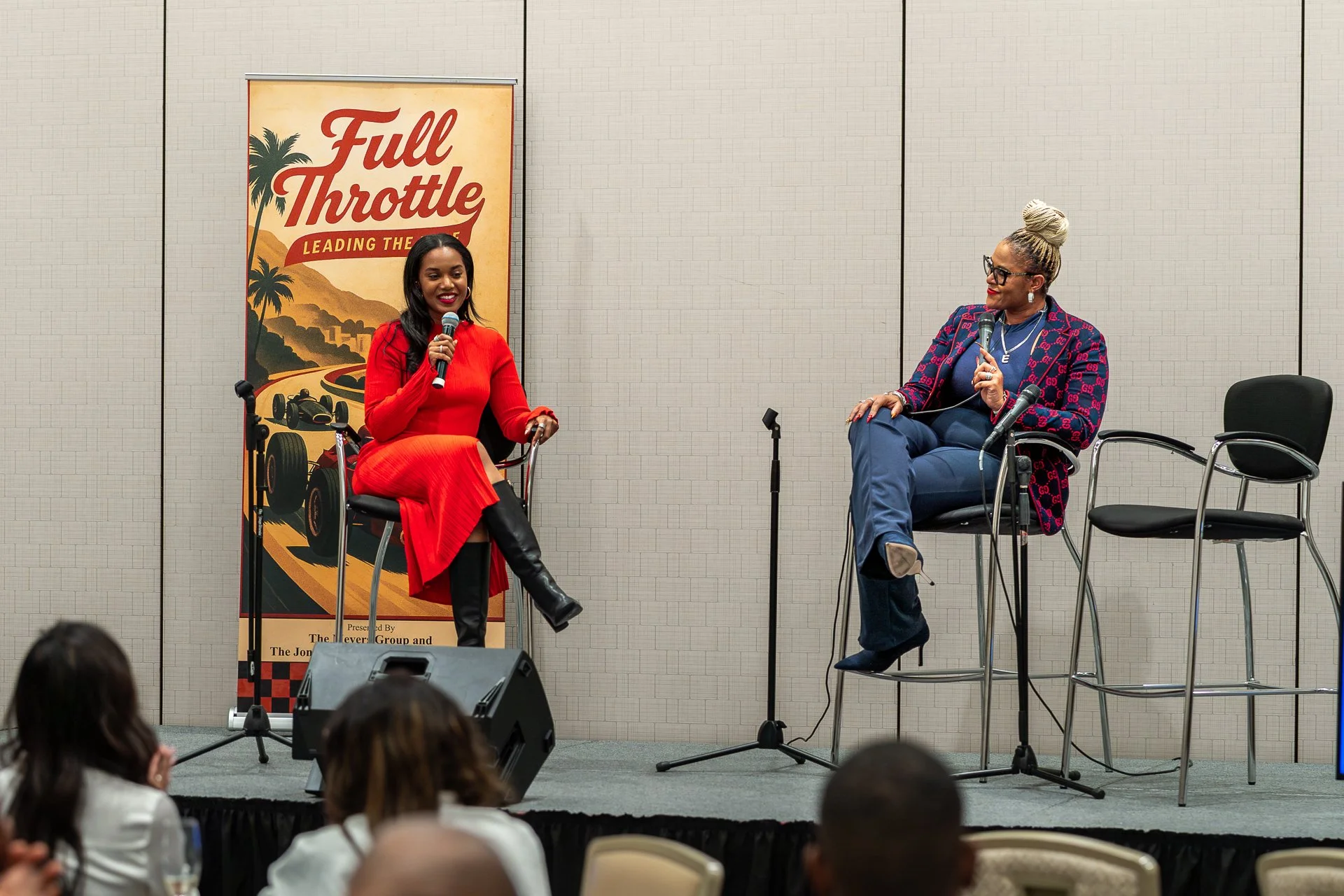 Two women on stage engaging in a discussion, one in a red dress and black boots, the other in a patterned blazer and blue pants, holding microphones. A banner behind them reads "Full Throttle: Leading the Race," with racing imagery.