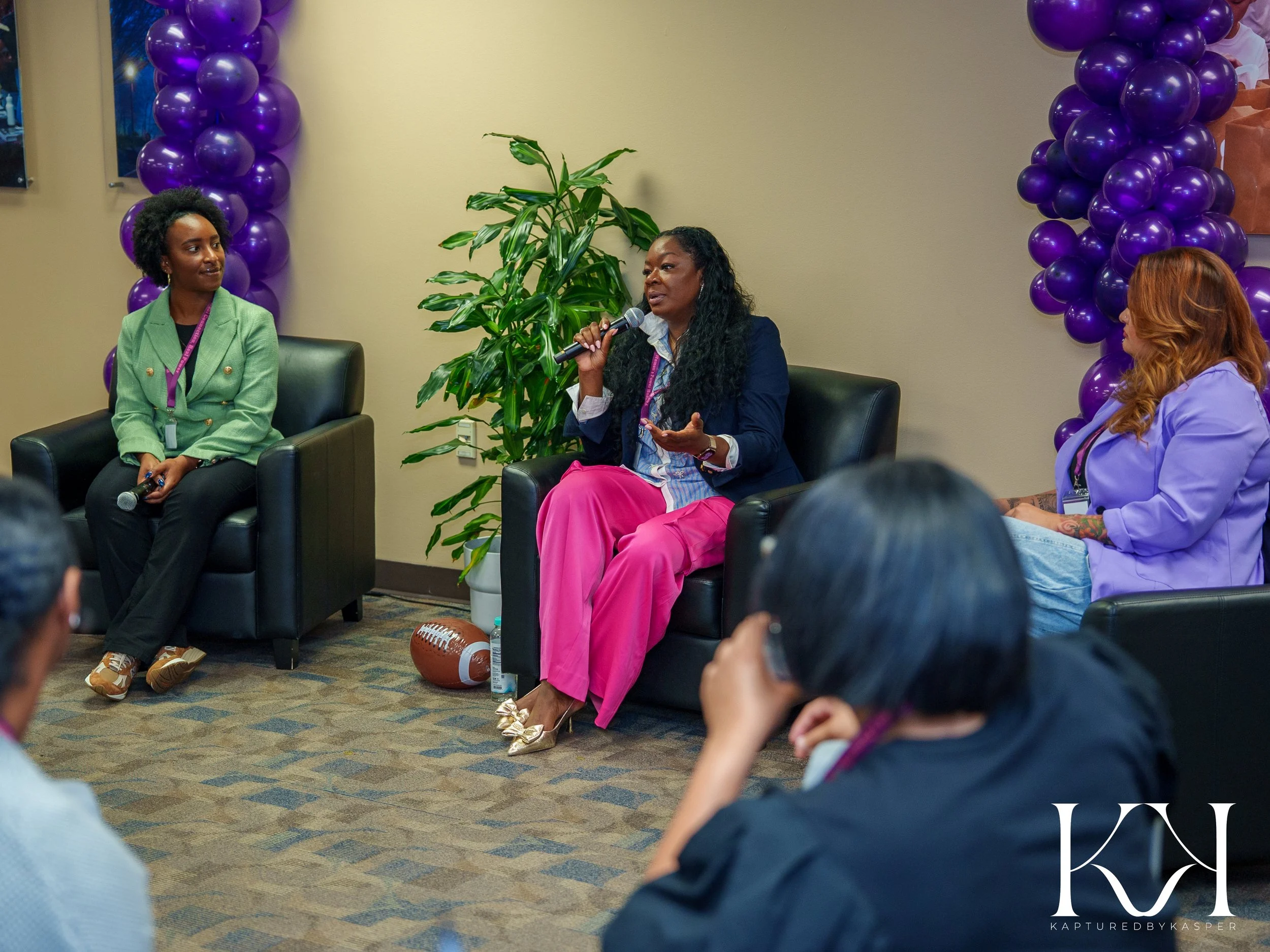 Three women are sitting in black armchairs in a room decorated with purple balloons. The woman in the middle is speaking into a microphone, wearing a dark blazer and pink pants. The woman on the left is wearing a green blazer, and the woman on the right has red hair, wearing a light purple blazer. There is a potted plant and a football on the floor.