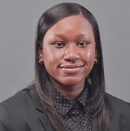 A professional portrait of a young Black woman with straight black hair, wearing a black blazer over a polka dot shirt, against a gray background.