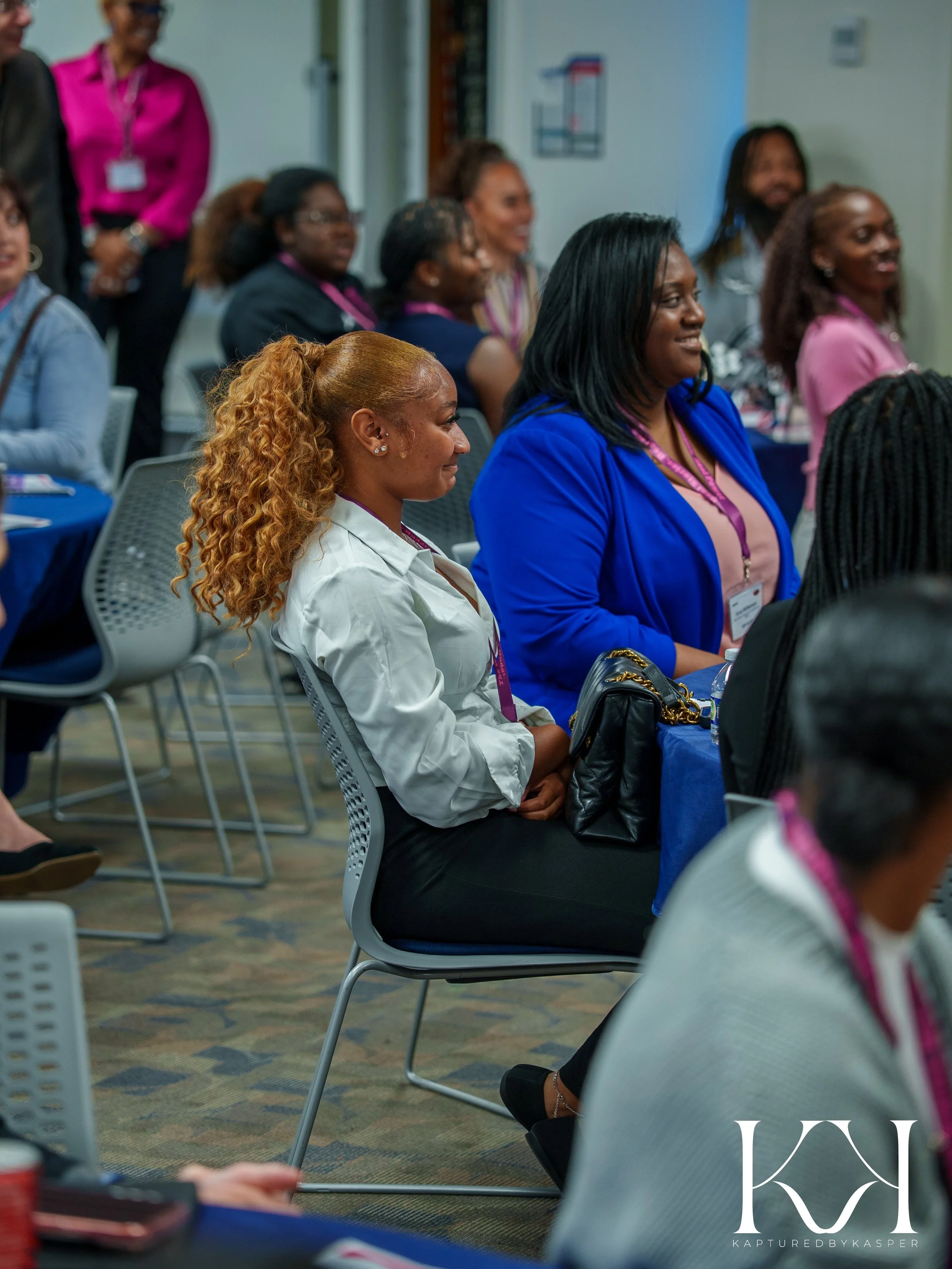Group of women attending a conference or seminar, seated at tables, listening attentively.