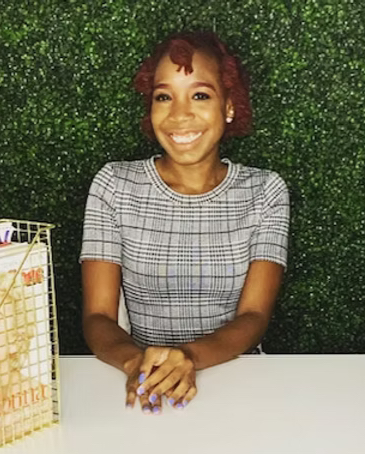Smiling woman with short red hair and ear gauges, sitting at a table with a green leafy background.