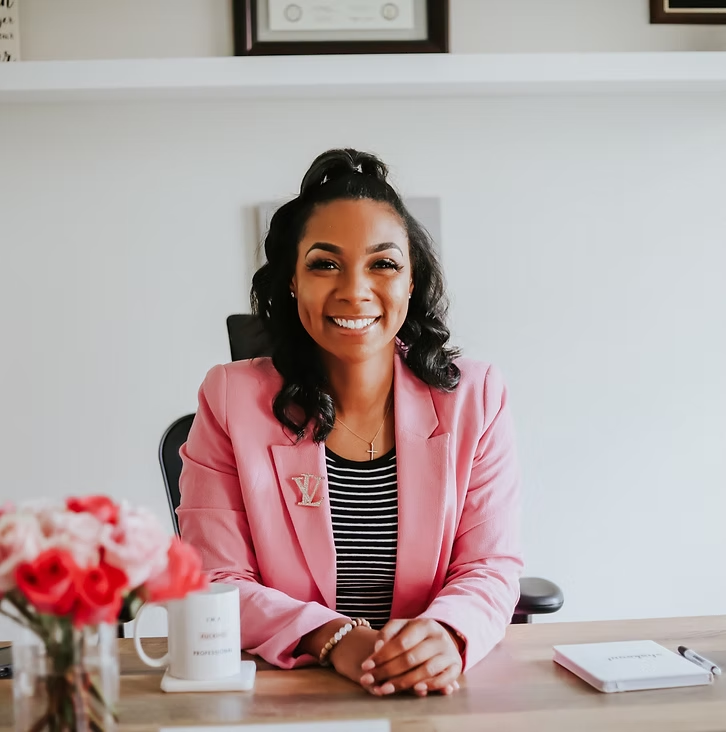 Smiling woman sitting at desk in office with pink blazer, striped shirt, and flowers on desk.