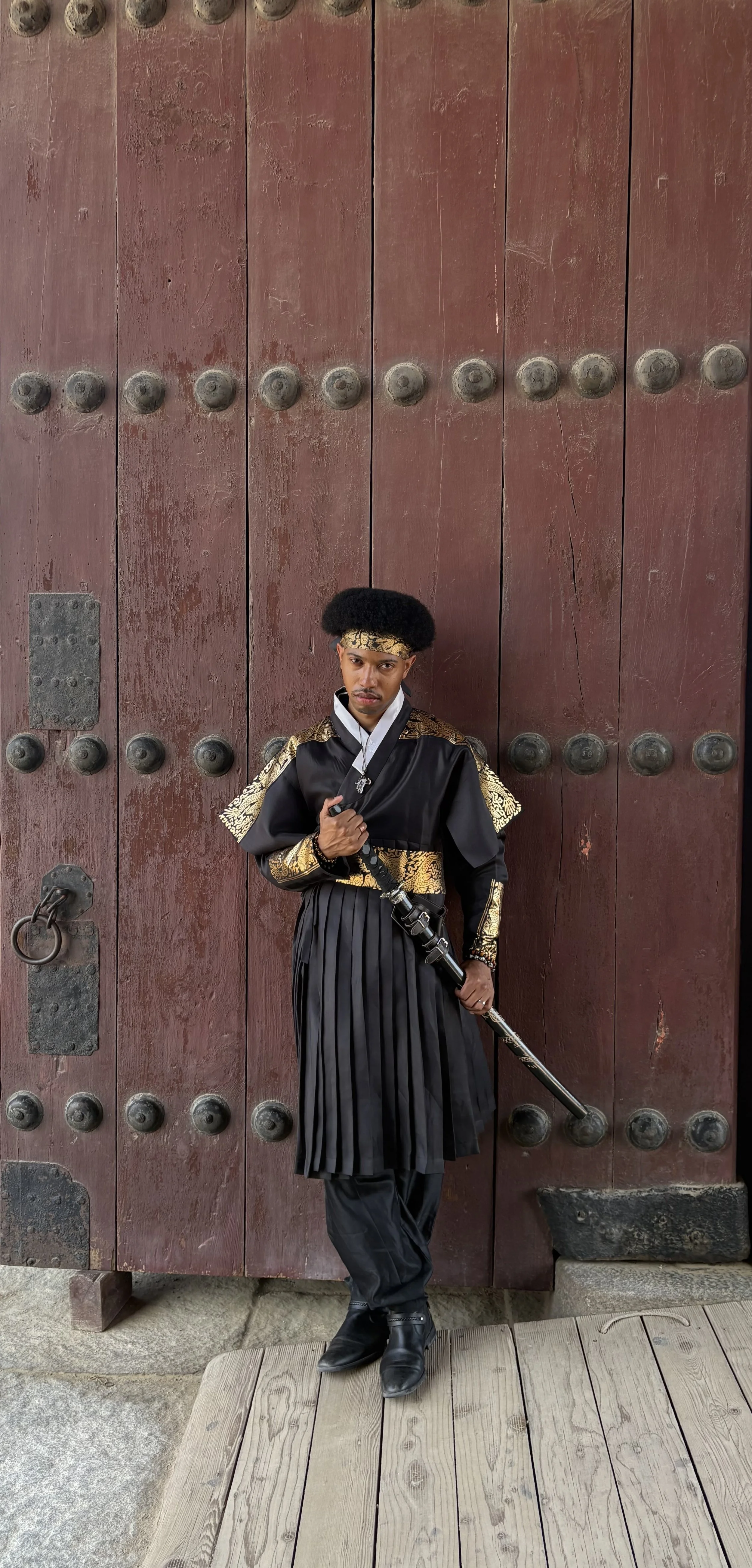 A man dressed in traditional armor standing in front of a large wooden door with metal studs, holding a sword.
