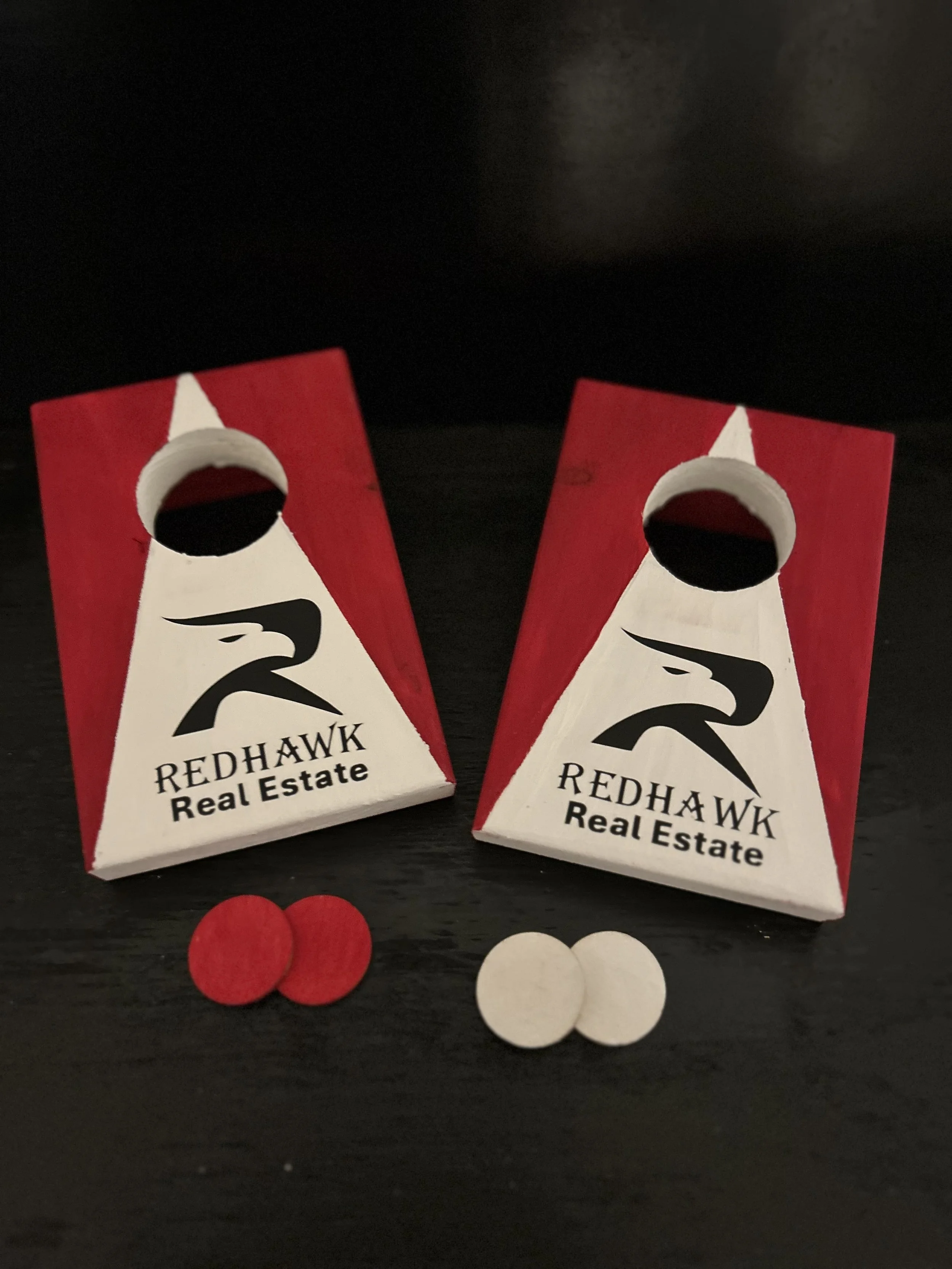 Two small red and white boxes with the logo and text 'Redhawk Real Estate'. There are three red and three white circular tokens below the boxes on a black surface.