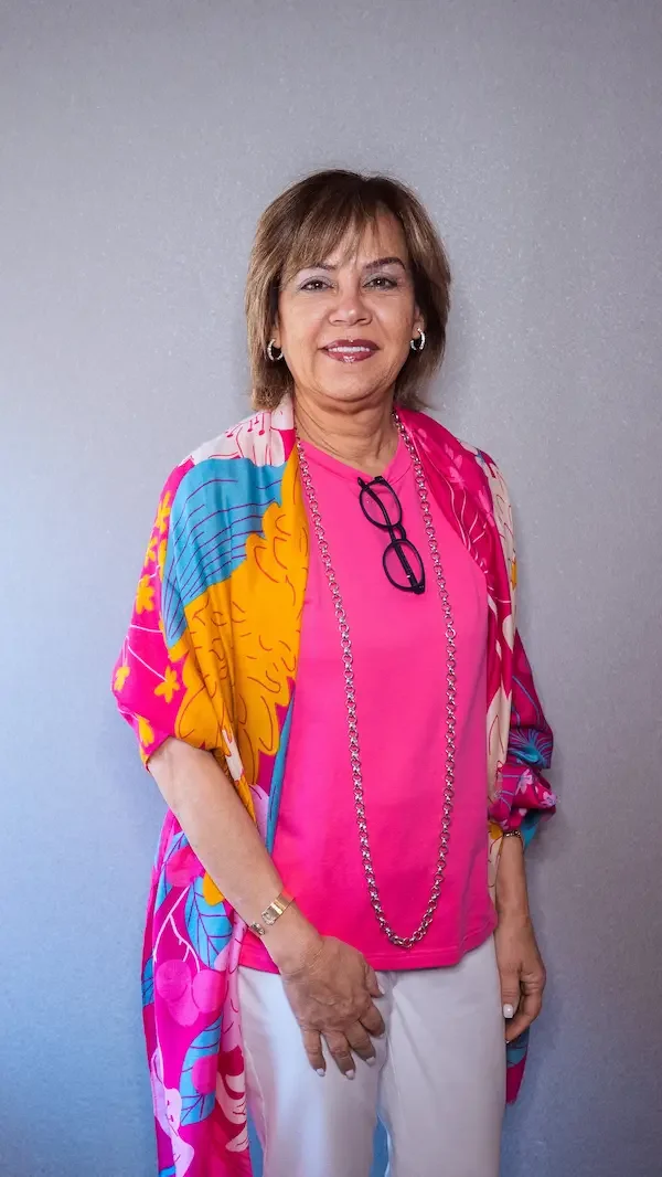 Mujer adulta con cabello castaño, usa gafas colgando del cuello, vestido rosa y pantalones blancos, lleva una bufanda o vestido de abrigo con estampado colorido de flores, en un fondo gris.