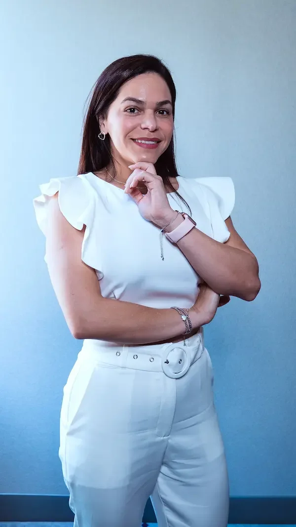 Mujer con blusa blanca y pantalones blancos posa con una mano en la cara y la otra cruzada, sonriendo