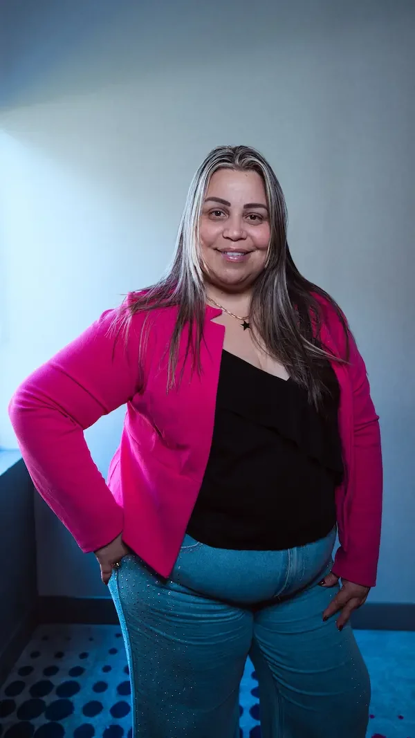 Mujer con cabello rubio y castaño, usando chaqueta rosa y pantalones azul claro, posando en interior con luz natural y fondo gris.