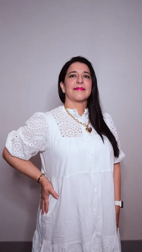 Mujer de cabello largo y oscuro, vestida con blusa blanca, con una mano en la cadera, usando un collar de corazón dorado y reloj blanco, posando frente a una pared gris.