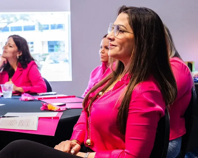 Mujeres en una reunión, vestidas con blusas rosadas, sentadas en una mesa con documentos y objetos decorativos.