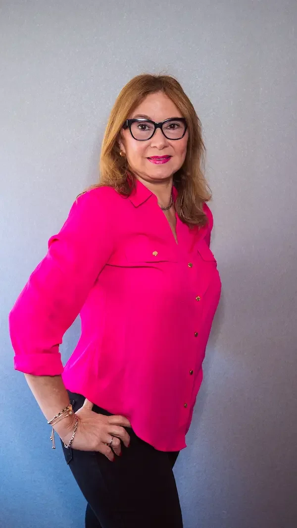 Mujer con gafas, camiseta rosa y pantalones negros, de pie junto a una pared gris, sonriendo y mirando a la cámara.