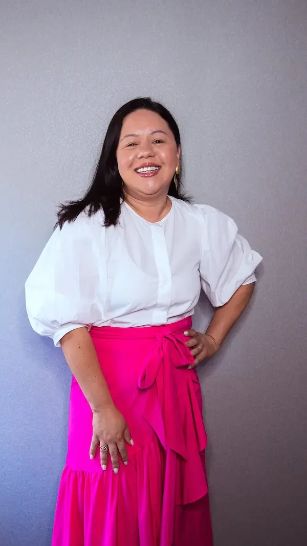 Mujer sonriendo con blusa blanca y falda larga rosa con lazo, de cabello oscuro y largo, posando contra pared gris.