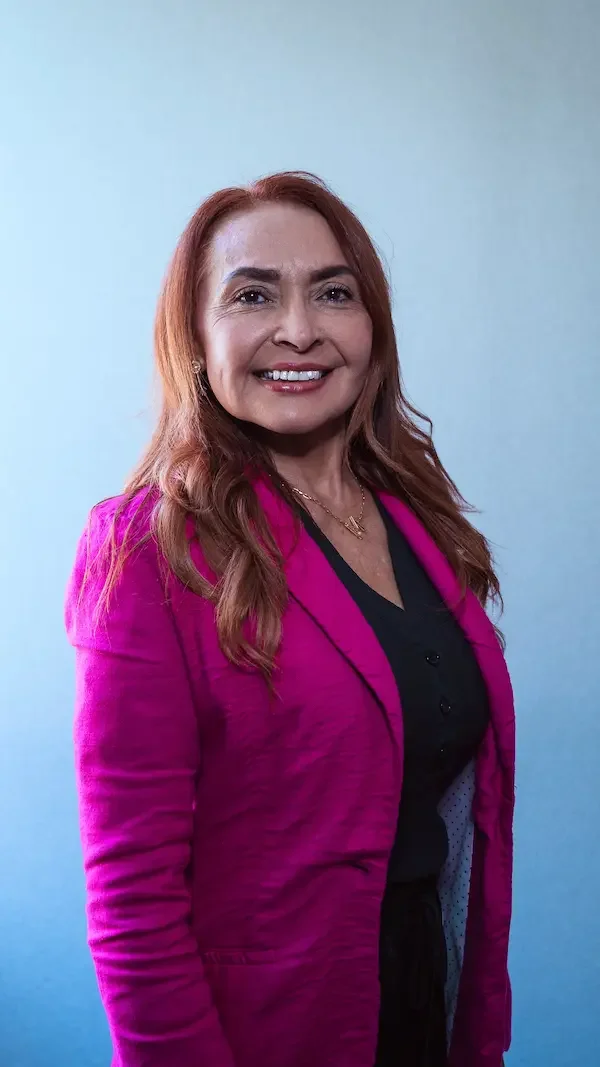 Retrato de una mujer con cabello rubio y cabello rizado, usando una chaqueta rosa y una camiseta negra, sonriendo frente a un fondo azul claro.