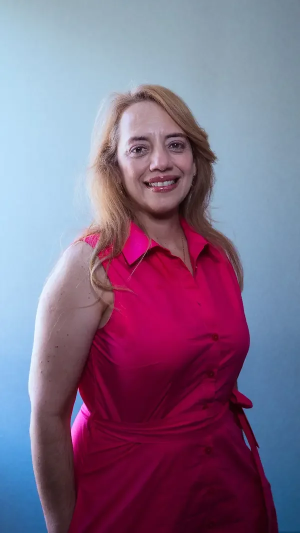 Mujer con cabello rubio, vistiendo blusa rosada, sonriendo, frente a fondo gris.