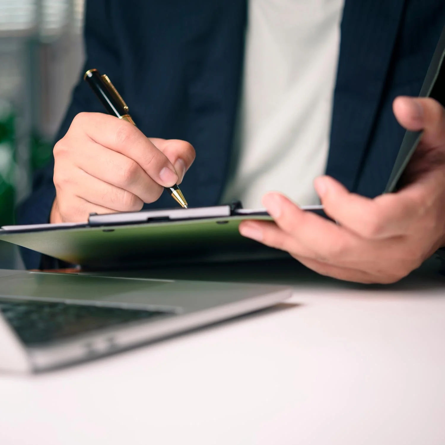 Person holding a pen and clipboard while reviewing documents at a desk with a laptop