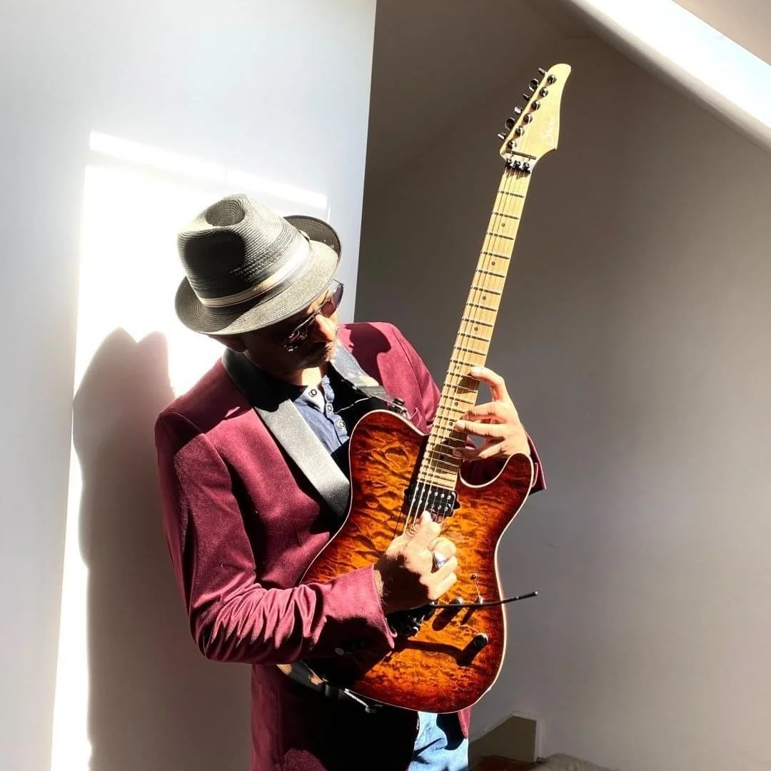 Danny John wearing a hat and sunglasses plays an electric guitar in a well-lit room.