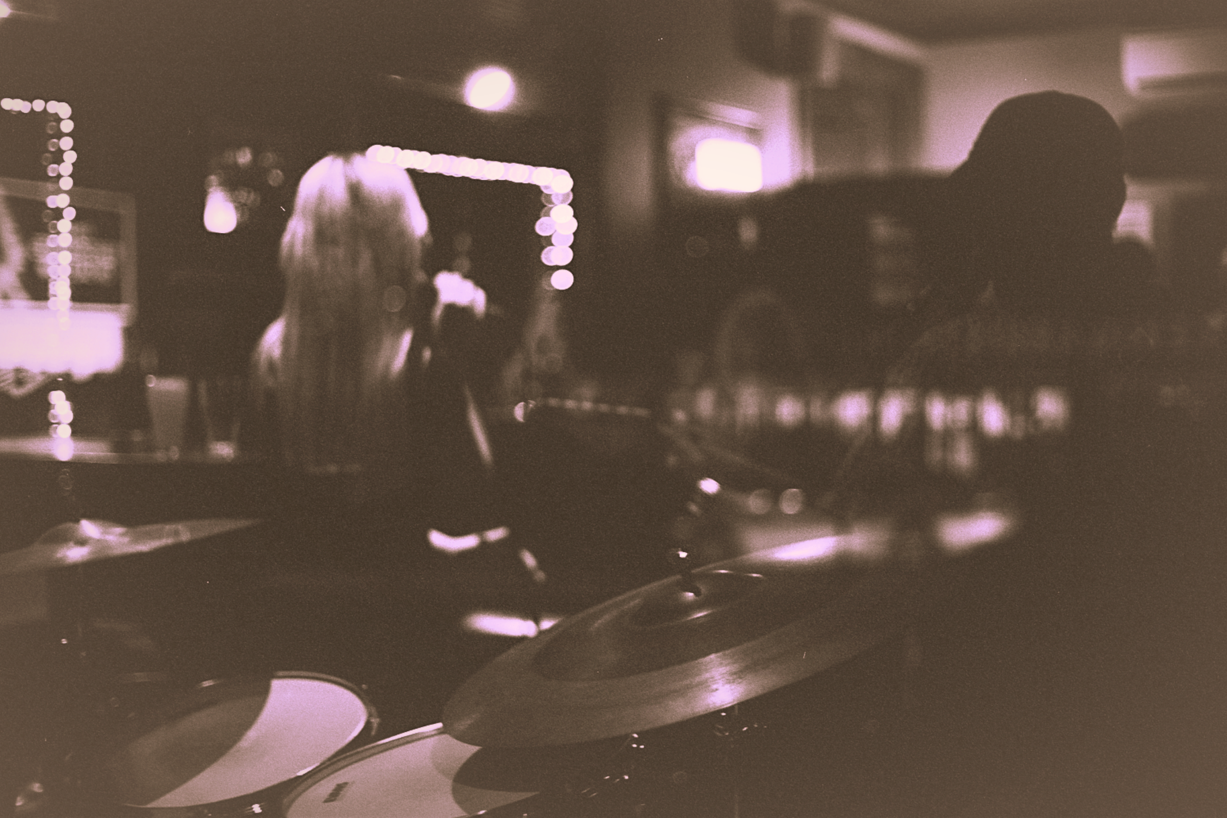 Vintage-style black and white image of Orla Birkett performing in a dimly lit venue with a visible drum set and blurred silhouettes.