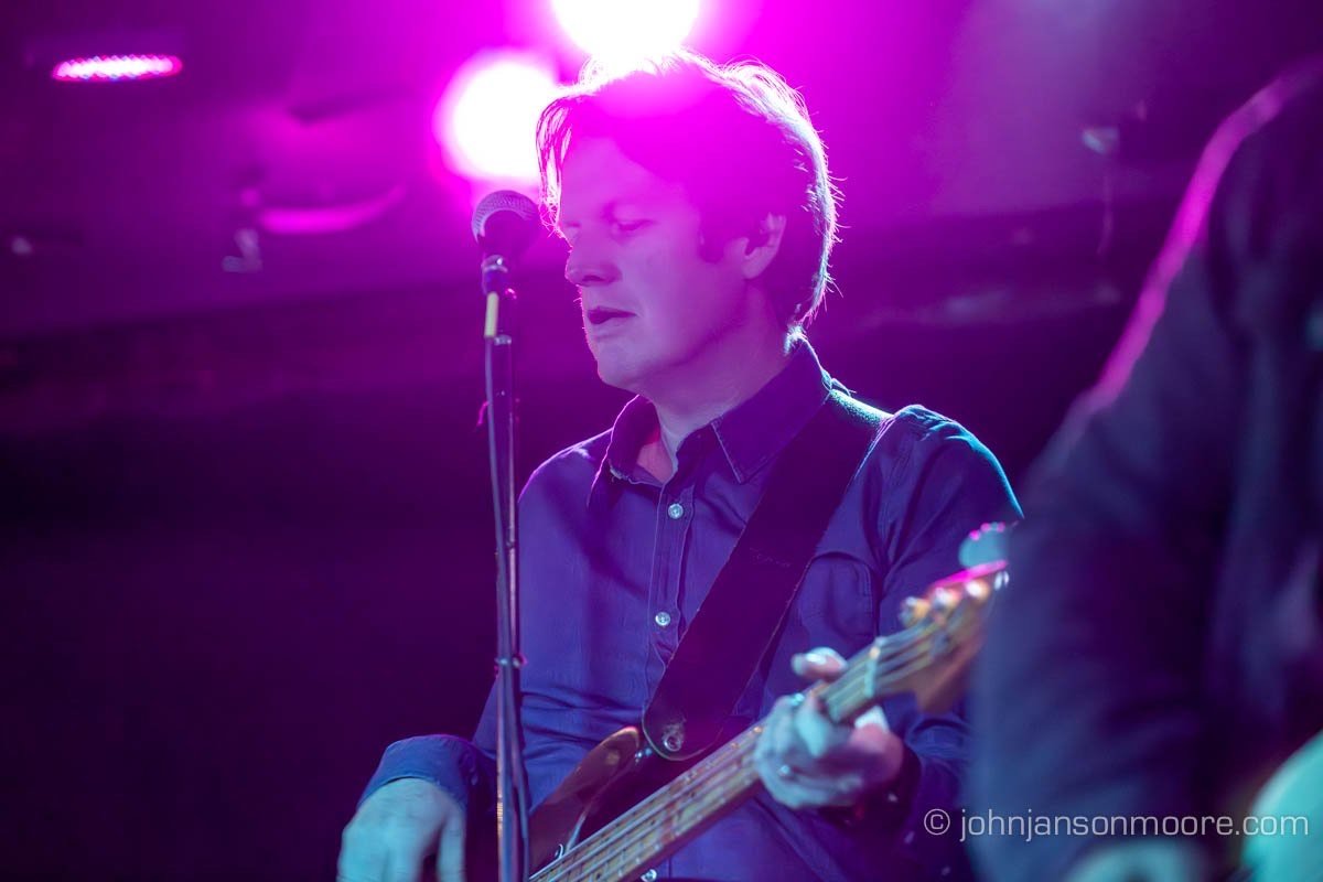 Dave Howell playing bass guitar on stage under purple lighting.