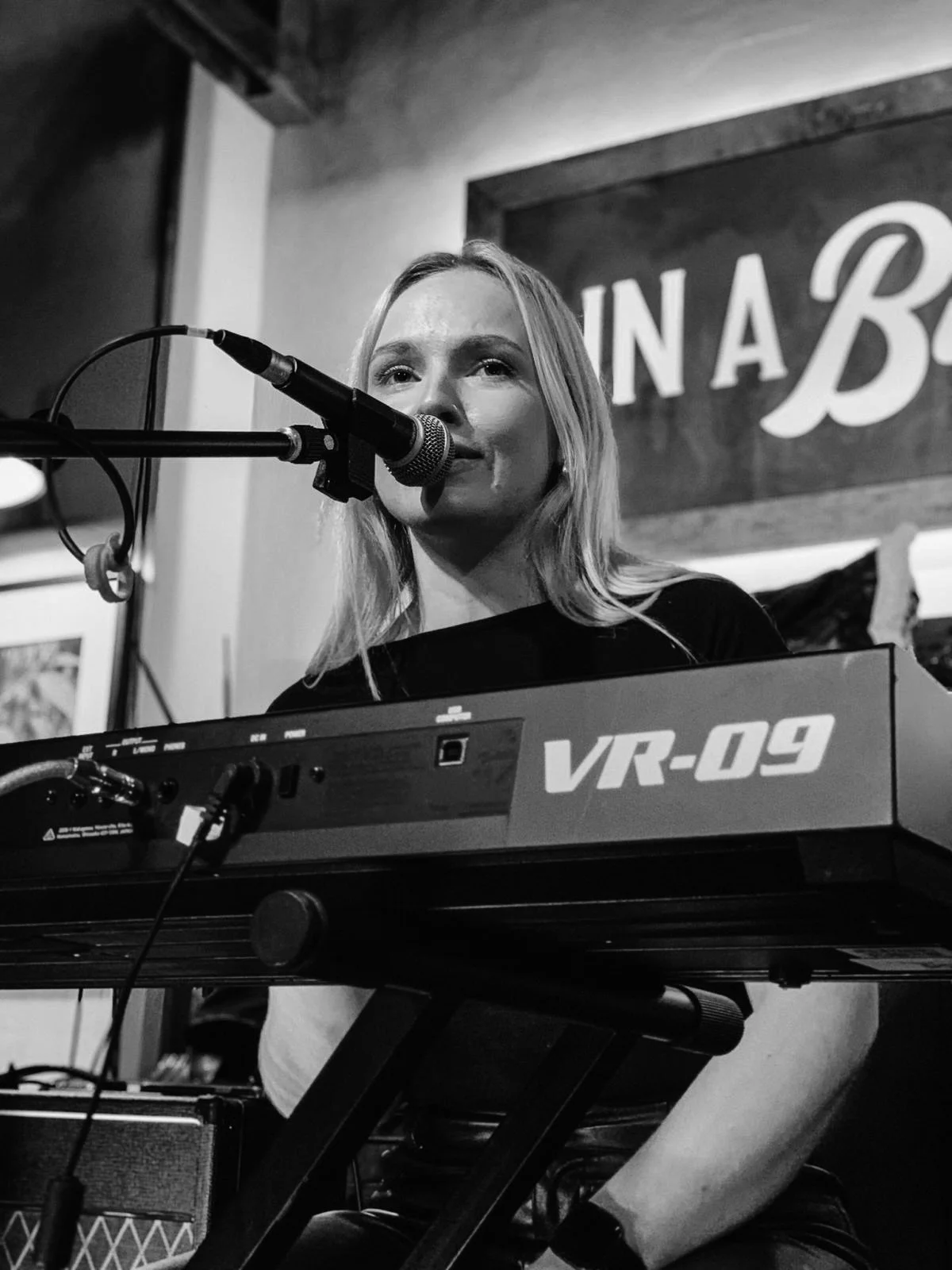 Black and white photo of Orla Birkett playing a VR-09 keyboard and singing into a microphone on stage.