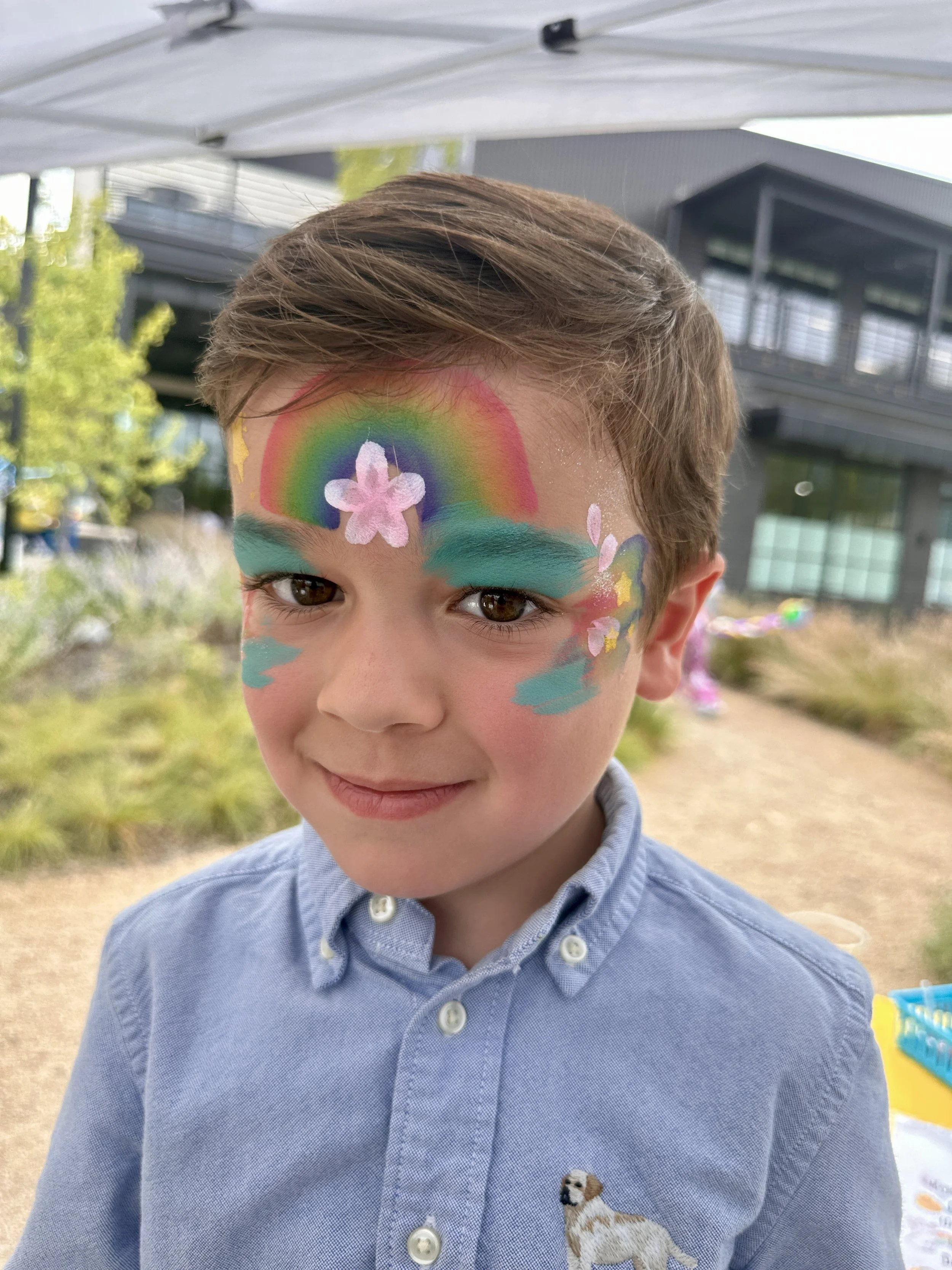 Young boy with colorful face paint including a rainbow, flowers, and clouds, smiling outdoors.