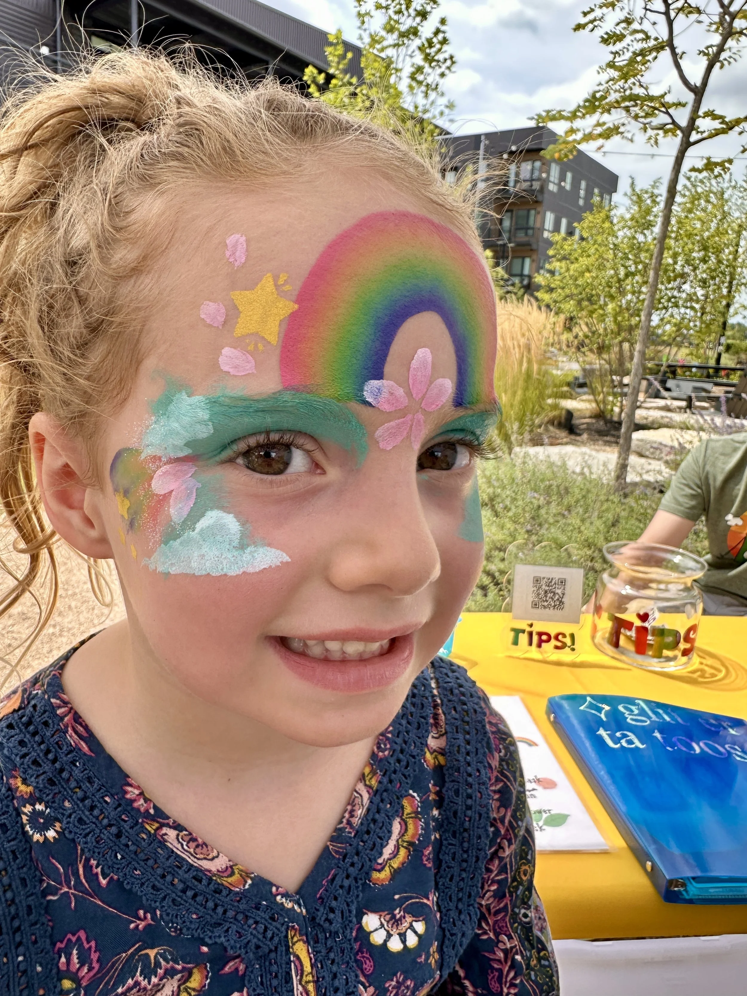 A young girl with face paint featuring a rainbow, clouds, pink flowers, and stars, smiling outdoors on a sunny day.