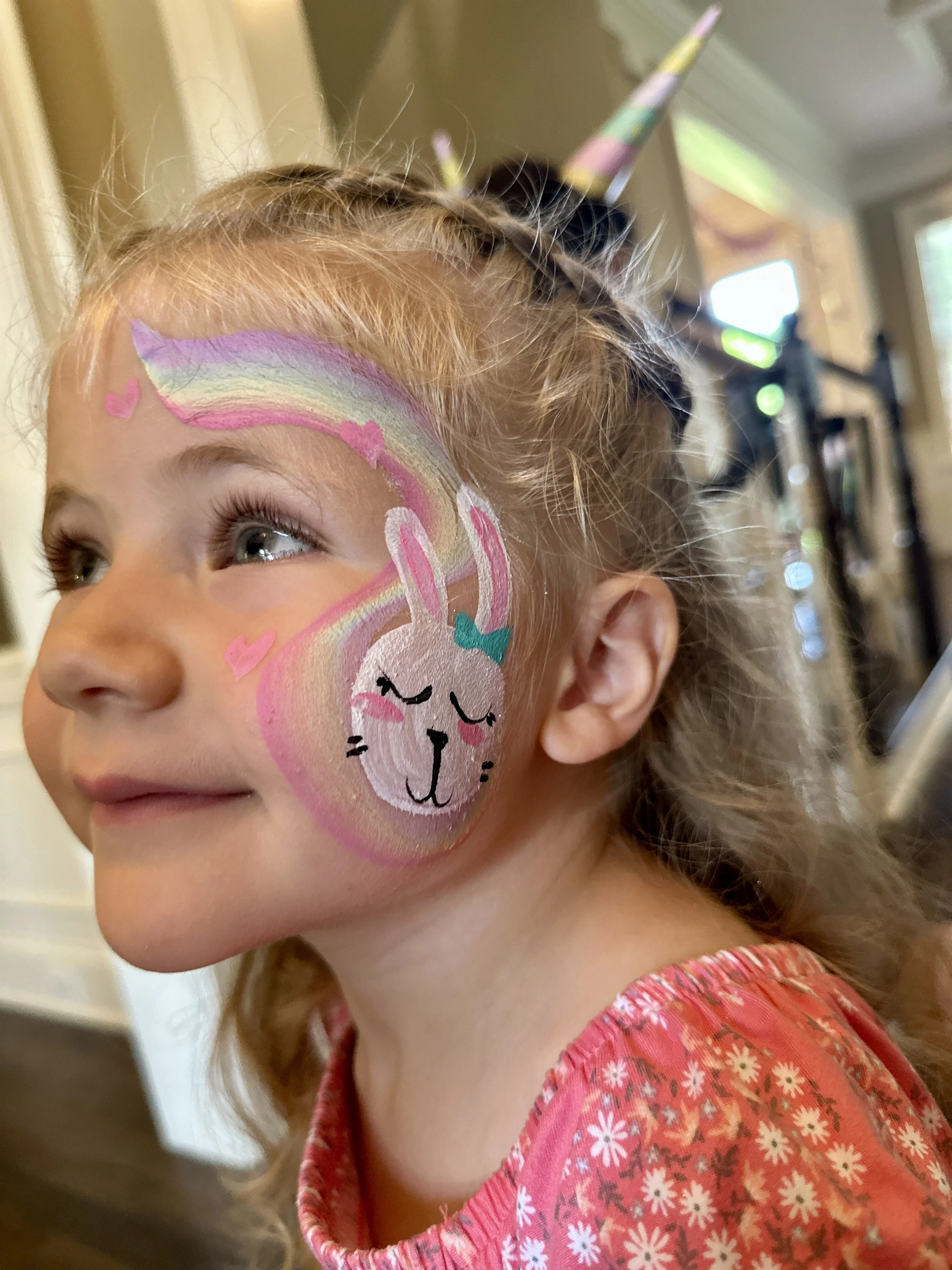 A young girl with face paint of a rainbow and a smiling bunny with closed eyes on her face. She is smiling and looking to her left, wearing a pink floral shirt. Her hair is styled with braids and a unicorn horn headband.