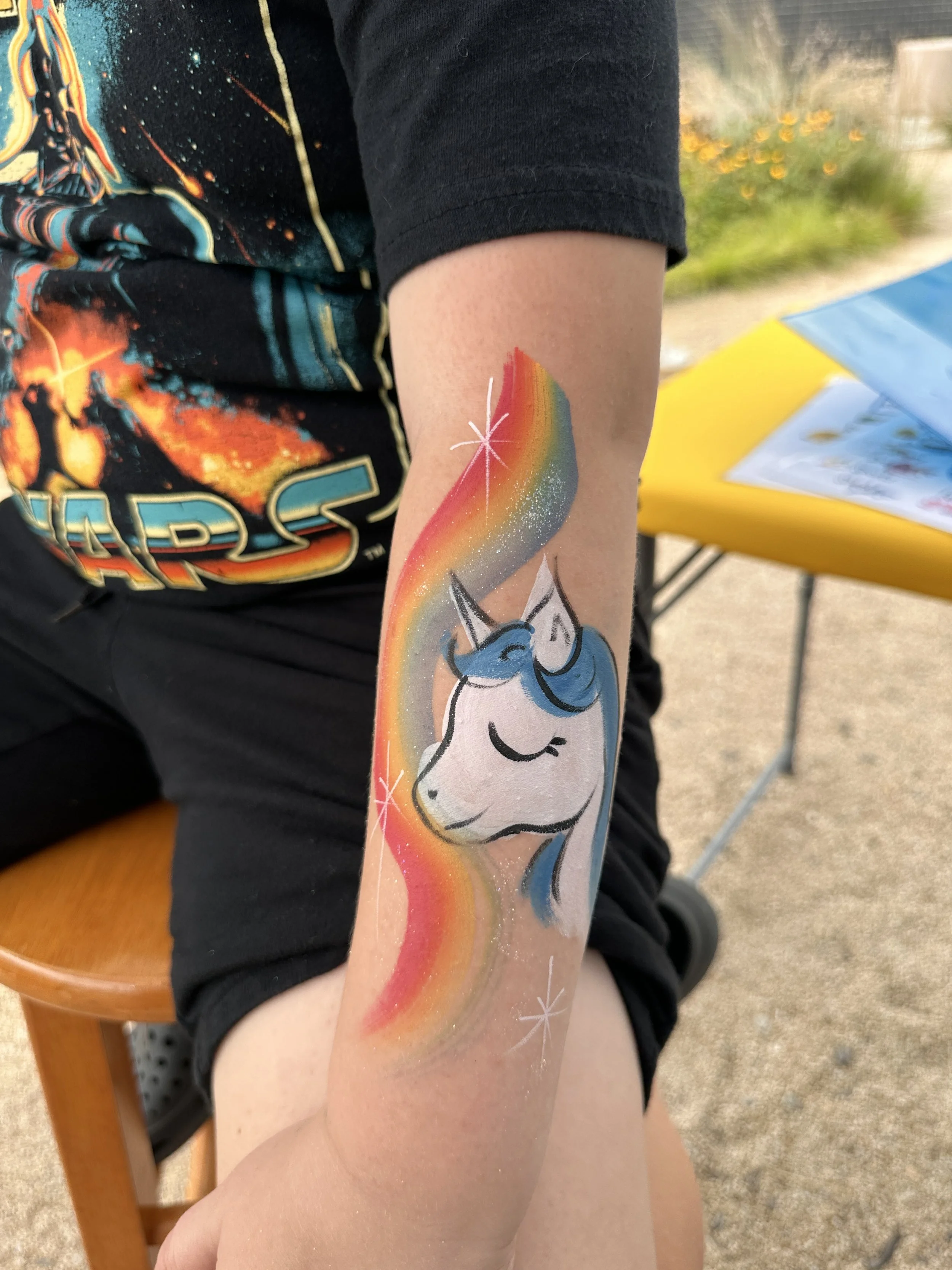 Colorful tattoo of a unicorn with a rainbow mane on a person's forearm, with a star and glitter accents.