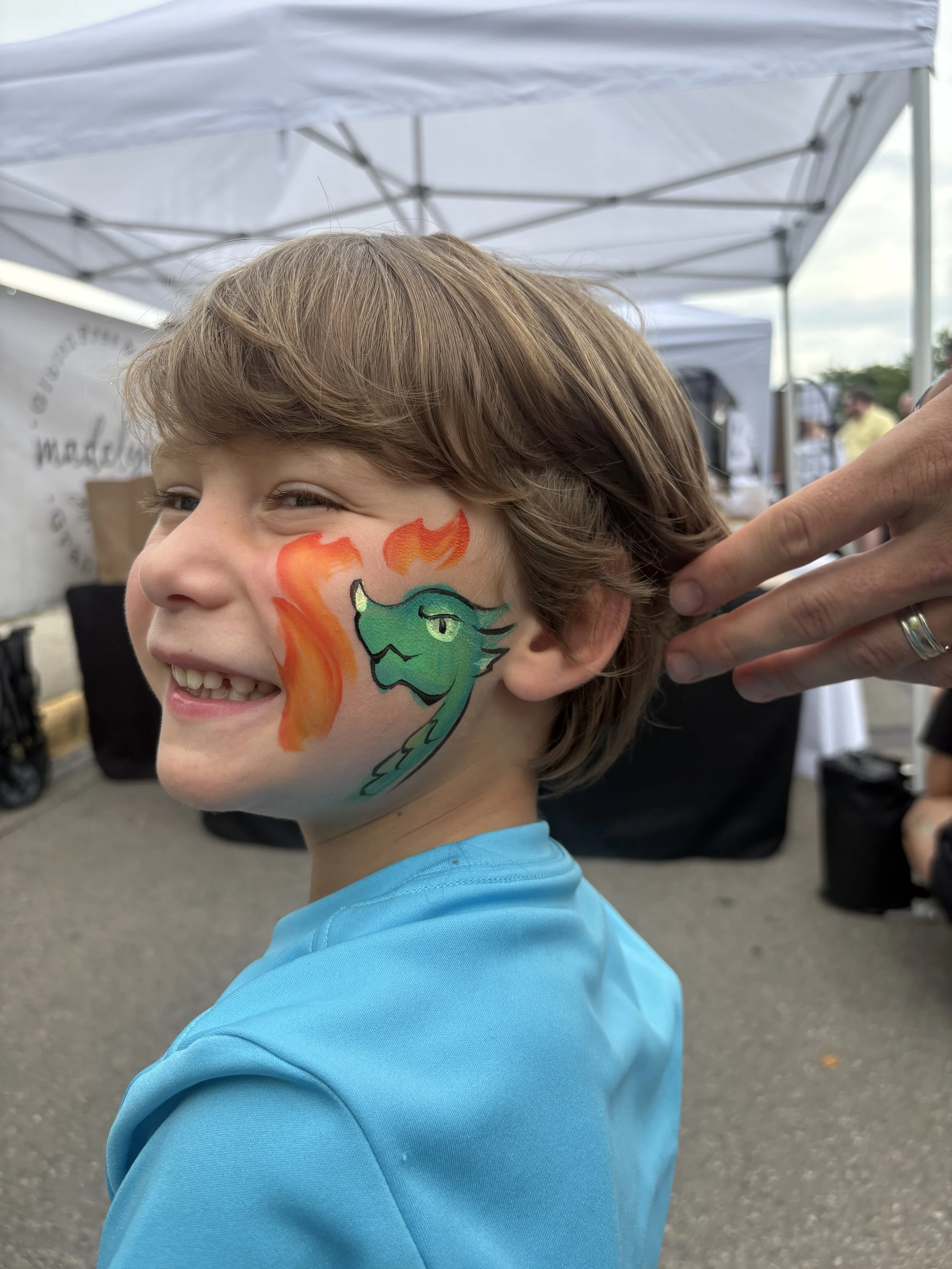 A young boy with a face painting of a green dragon breathing fire, smiling at an outdoor event.