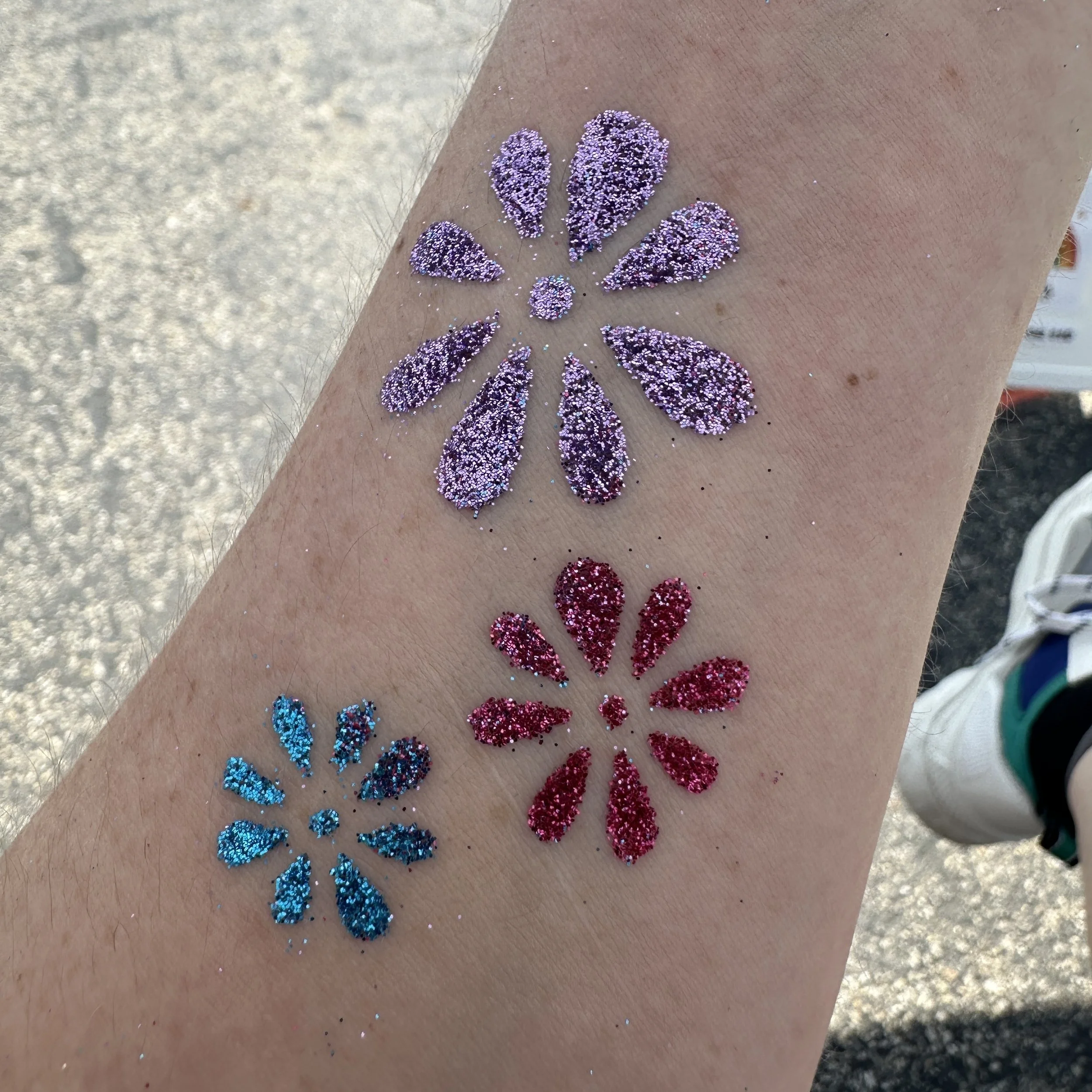 Arm with colorful glitter tattoo designs in purple, red, and blue arranged as flowers.