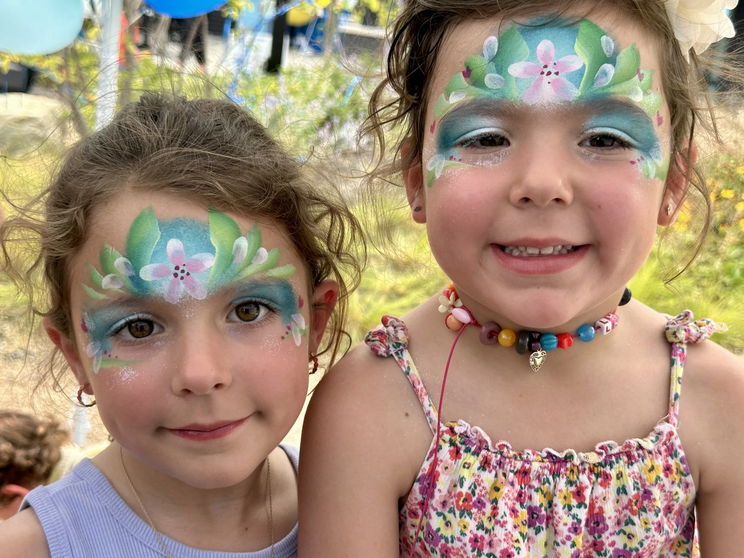 Two young girls with colorful face paint, featuring floral and leafy designs across their foreheads and eyes, standing outdoors at a festival or fair.