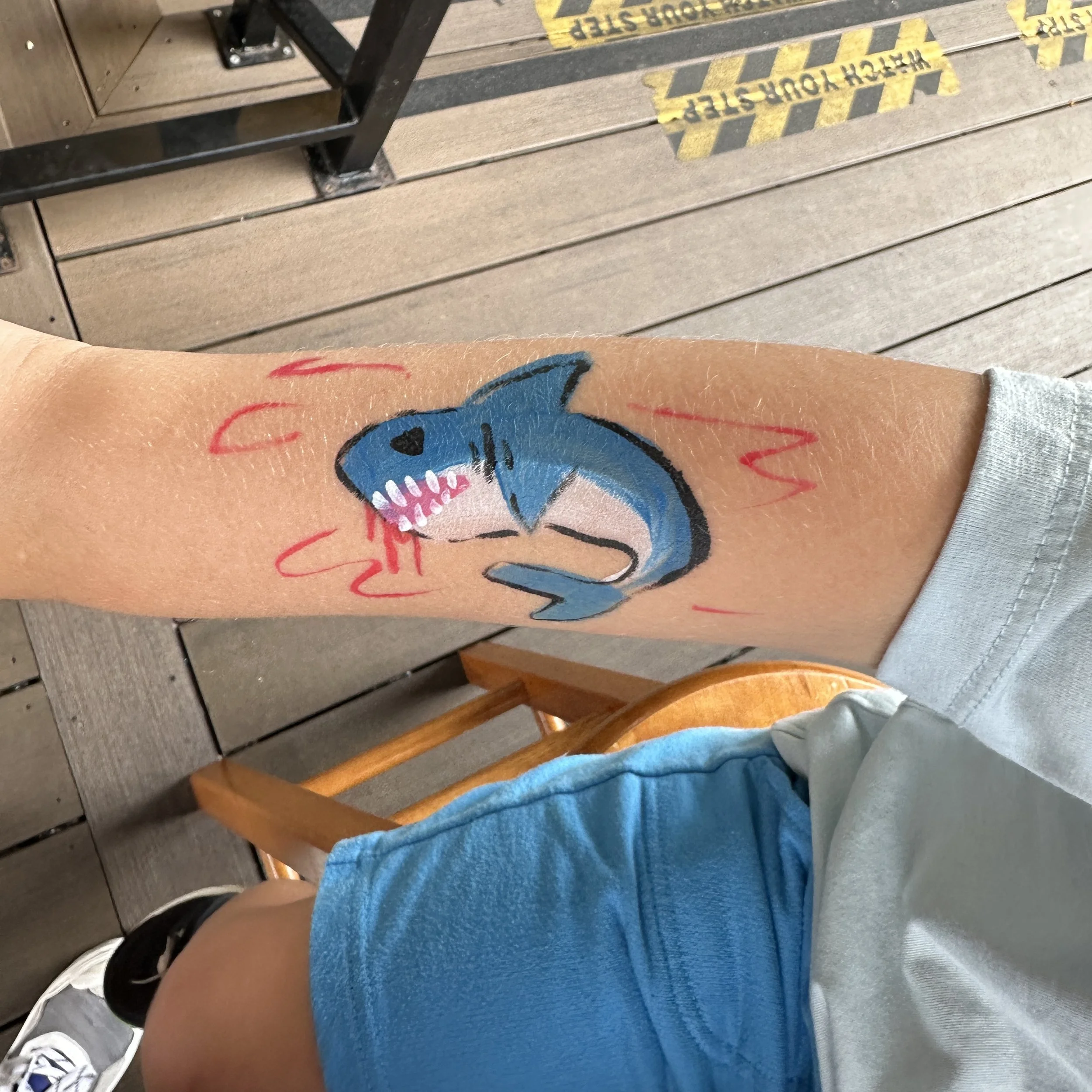 A person's arm with a painted tattoo of a blue shark with white teeth and black eyes, surrounded by red lines, on the forearm.