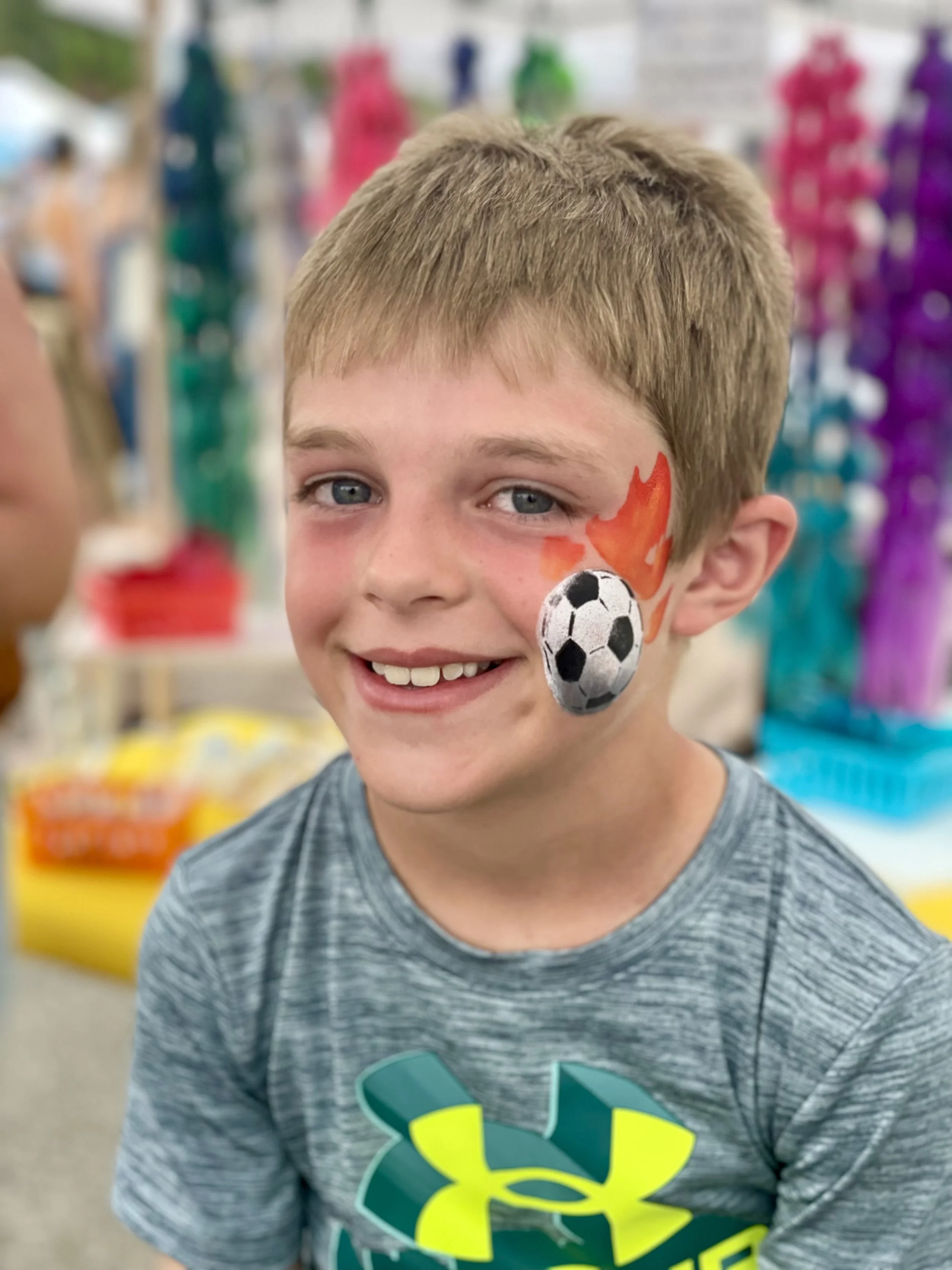 Young boy with face paint of a soccer ball and flames, smiling in a store with colorful merchandise in the background.