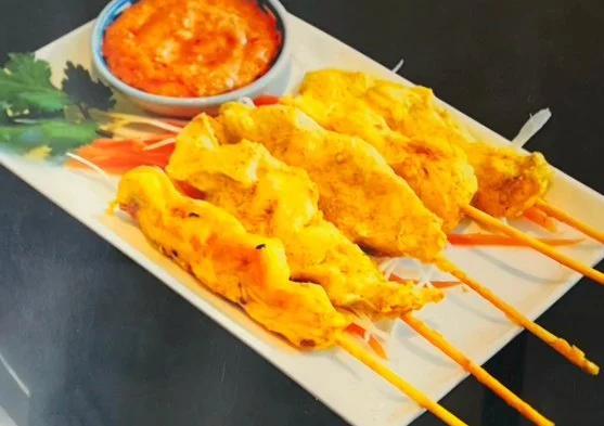 Four chicken satay skewers with a side of peanut satay dipping sauce on a rectangular white plate.