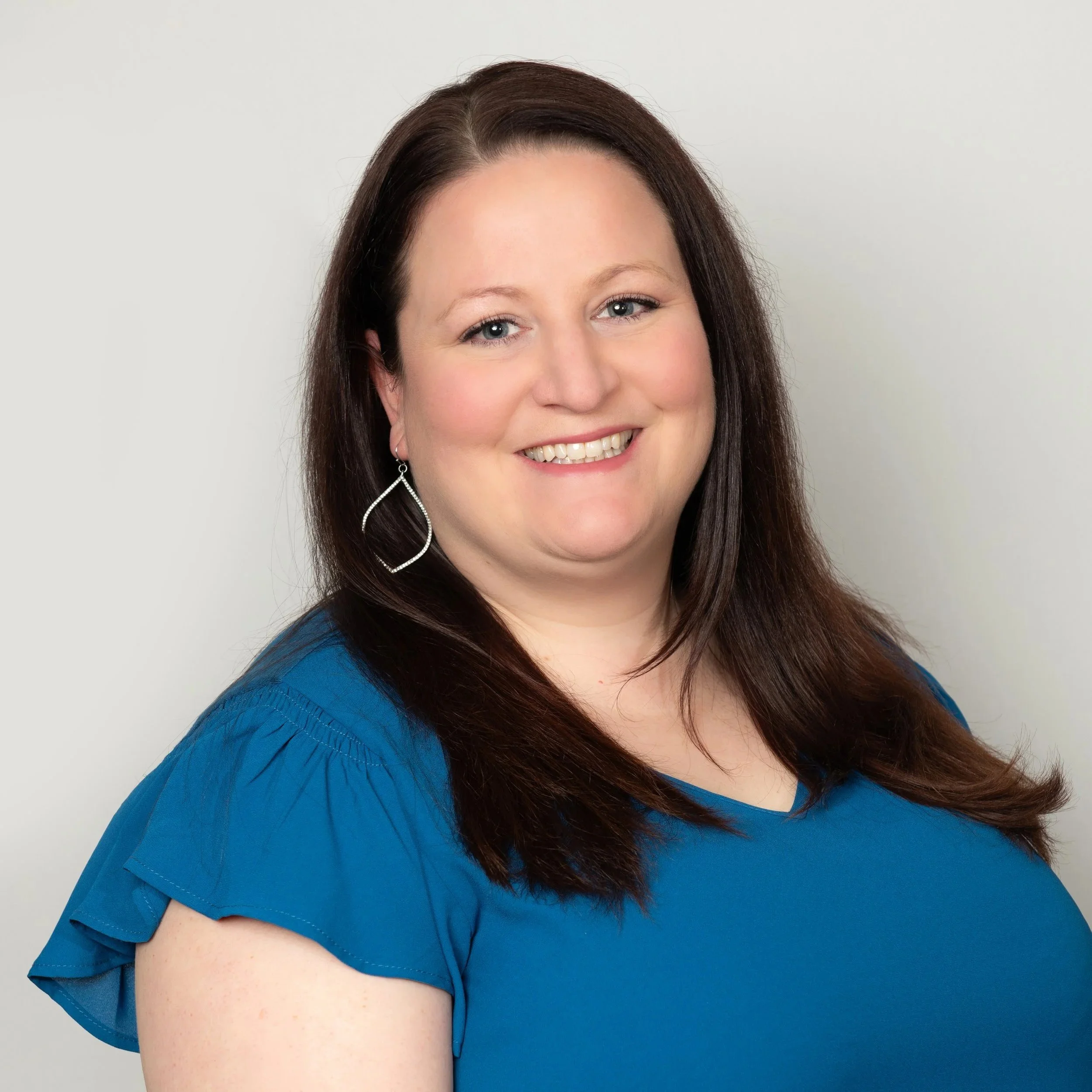A woman with long reddish-brown hair, wearing hoop earrings and a blue top, smiling outdoors with blurred trees in the background.