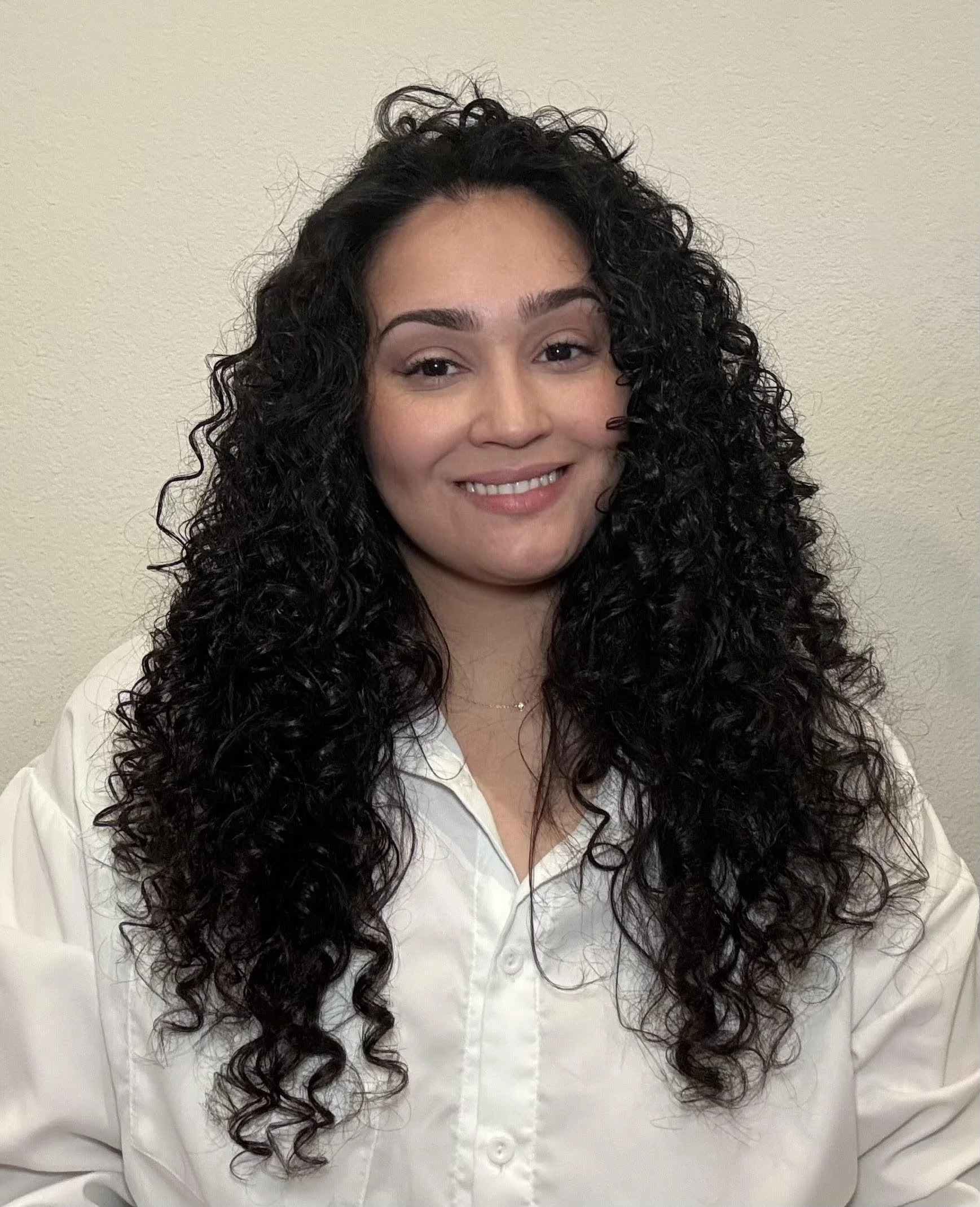 Headshot of a woman with long, wavy brown hair wearing a pink blouse, smiling softly against a neutral background.