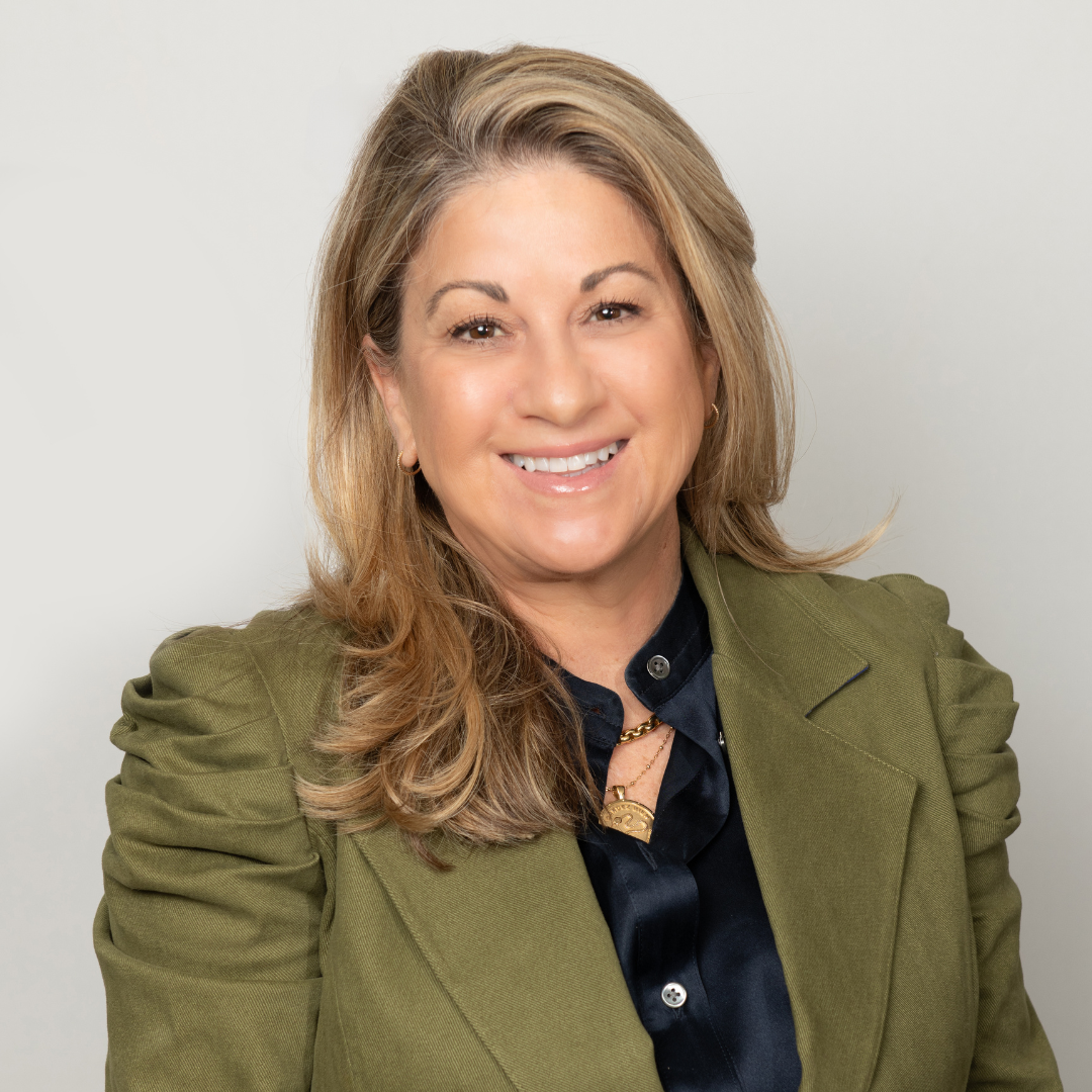 A woman with light skin and shoulder-length light brown hair styled in loose waves, smiling and wearing a green blazer over a black blouse, gold jewelry, and small earrings.