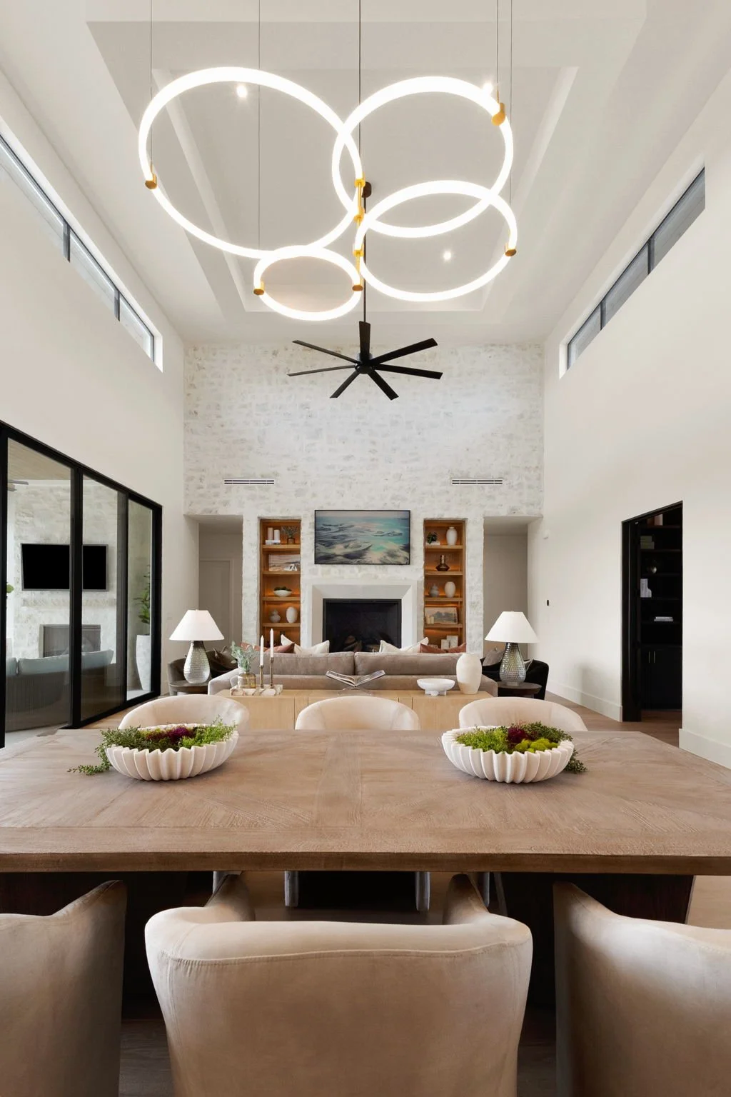 Modern living room with high ceiling, circular LED light fixture, black ceiling fan, white walls, beige furniture, fireplace with built-in shelving, large windows, and a dining table in the foreground.