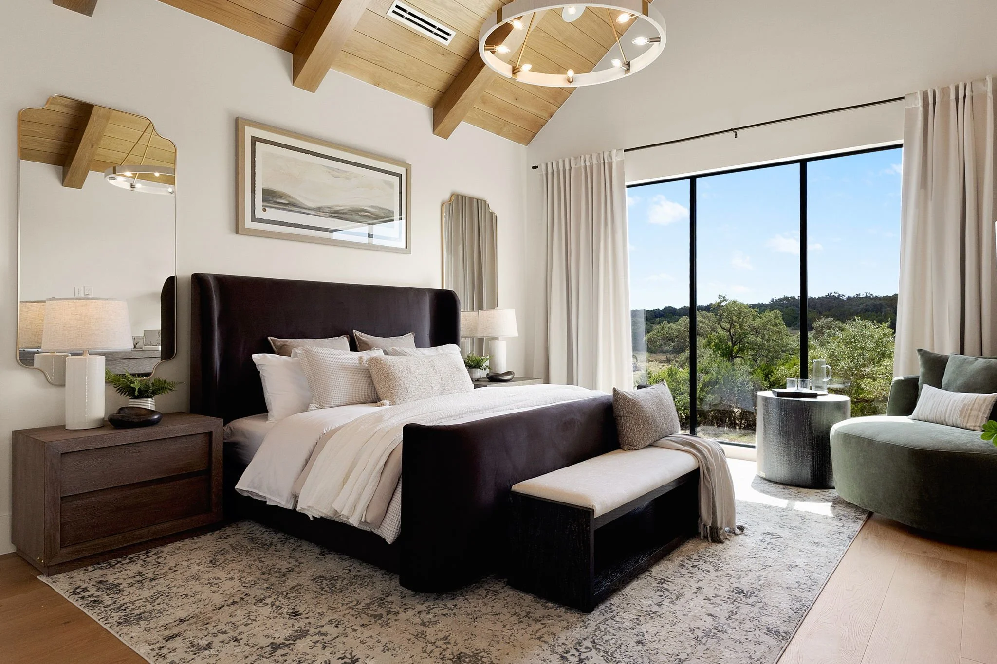 Modern bedroom with large glass window showing outdoor scenery, dark leather bed with white bedding, bedside tables with lamps, and sitting area with green armchair and small table.