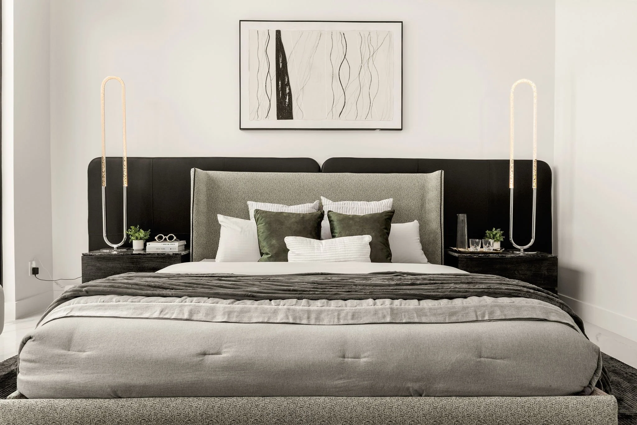 Modern bedroom with a large bed, gray and white bedding, black headboard, black nightstands, stylish lamps, and minimalist artwork on white walls.