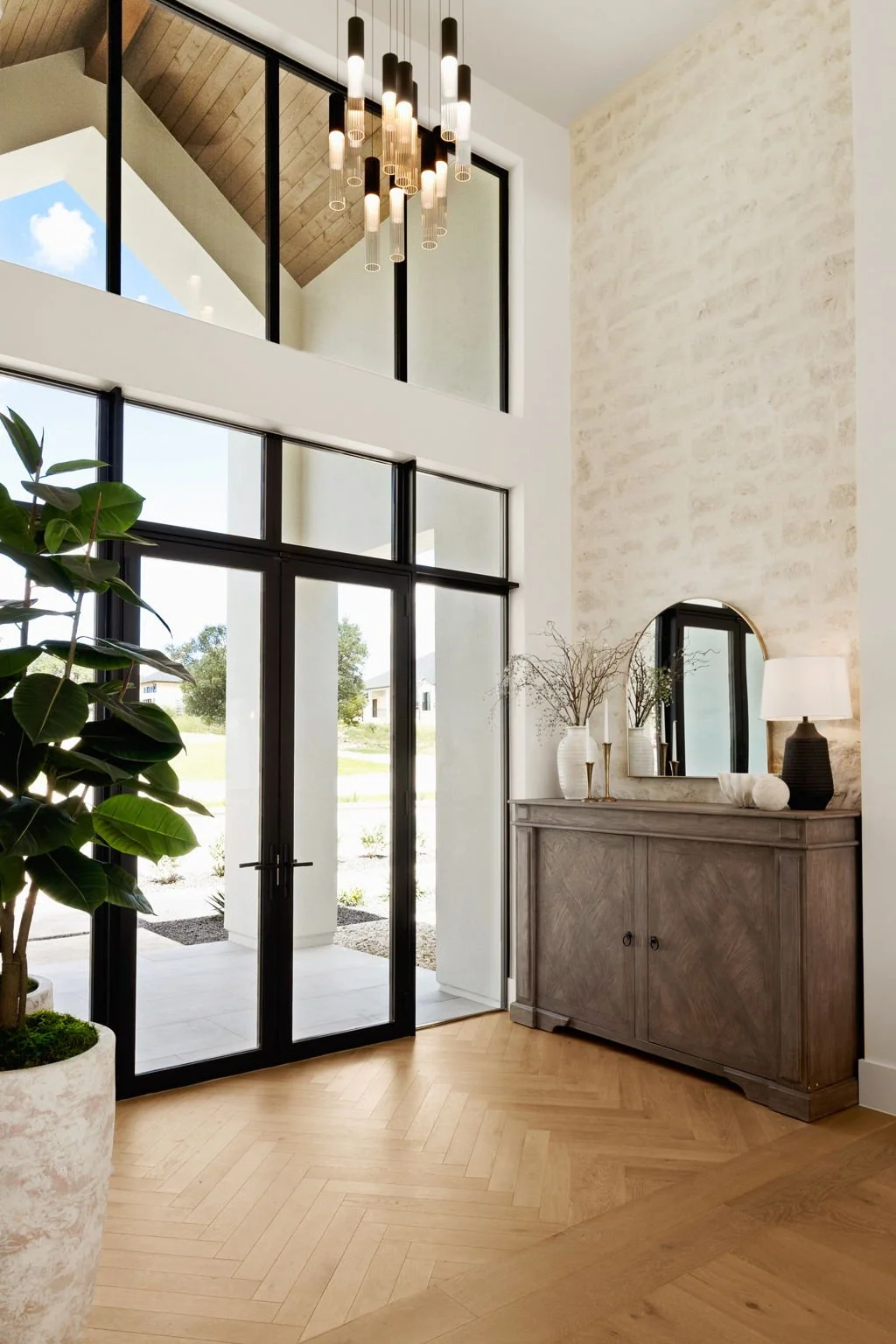 Modern home's entryway with large black-framed glass door, interior decor including a gray wooden sideboard with decorative vases and mirror, a tall potted plant, and a chandelier with multiple pendant lights hanging from the ceiling.