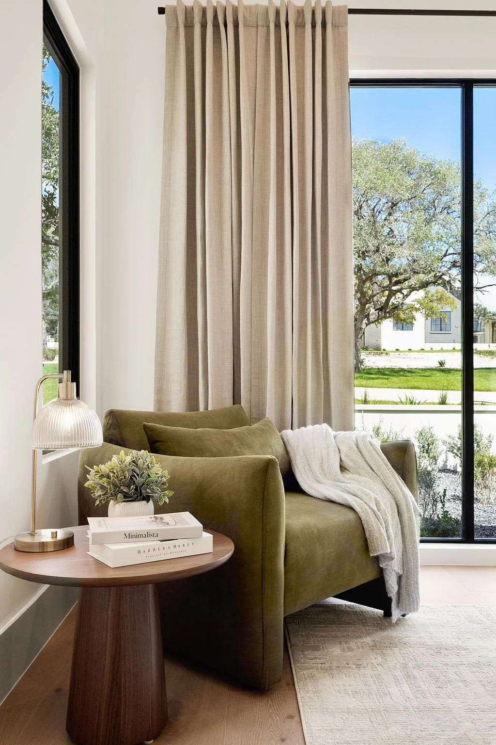 Living room with beige curtains, a green upholstered couch with a white throw blanket, a wooden side table with a small potted plant, books, and a modern table lamp. Large window shows a view of a tree and a house outside.