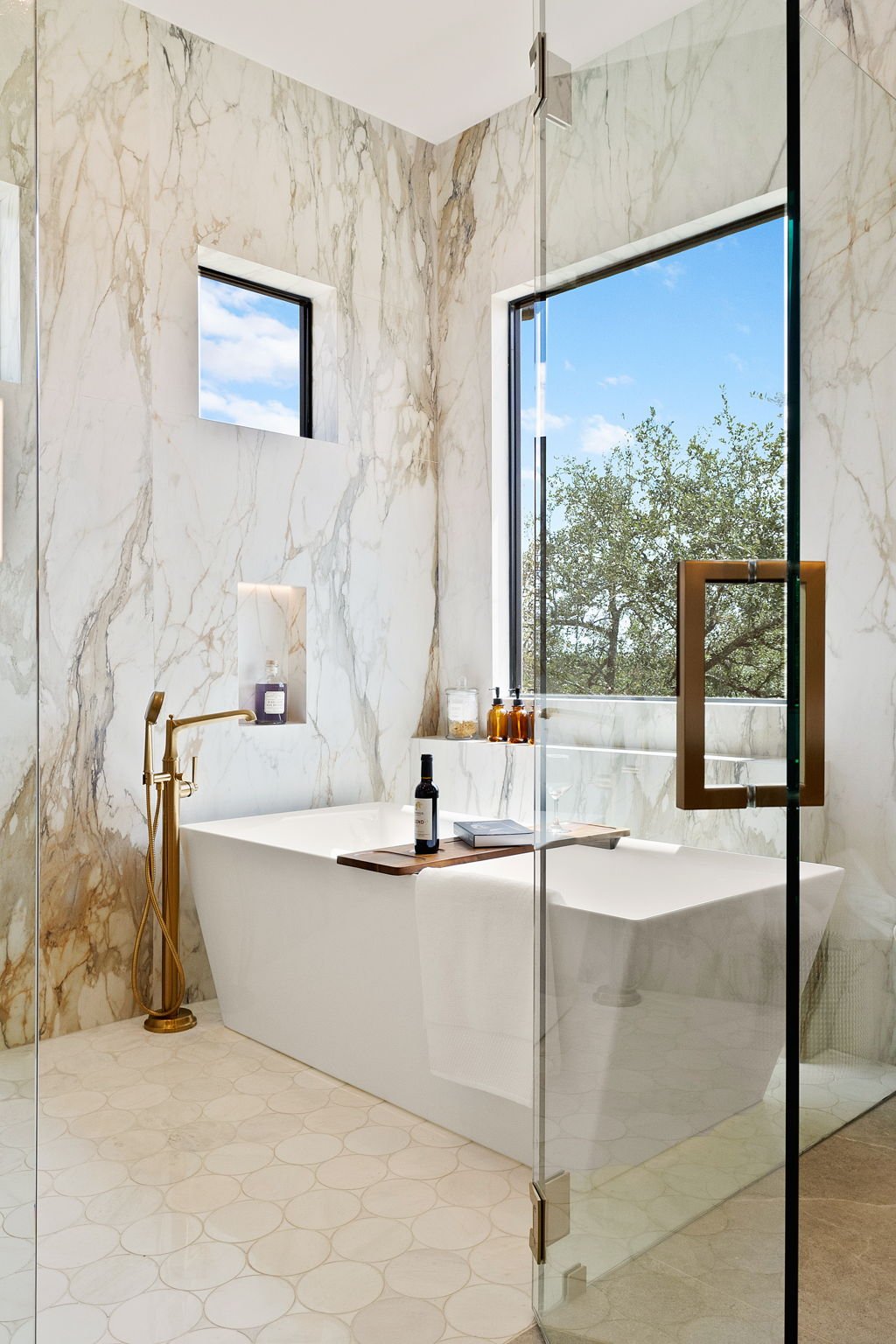 Modern bathroom with white marble walls and floor, a white bathtub, a large window showing outdoor trees and blue sky, and a glass shower door with a gold handle.
