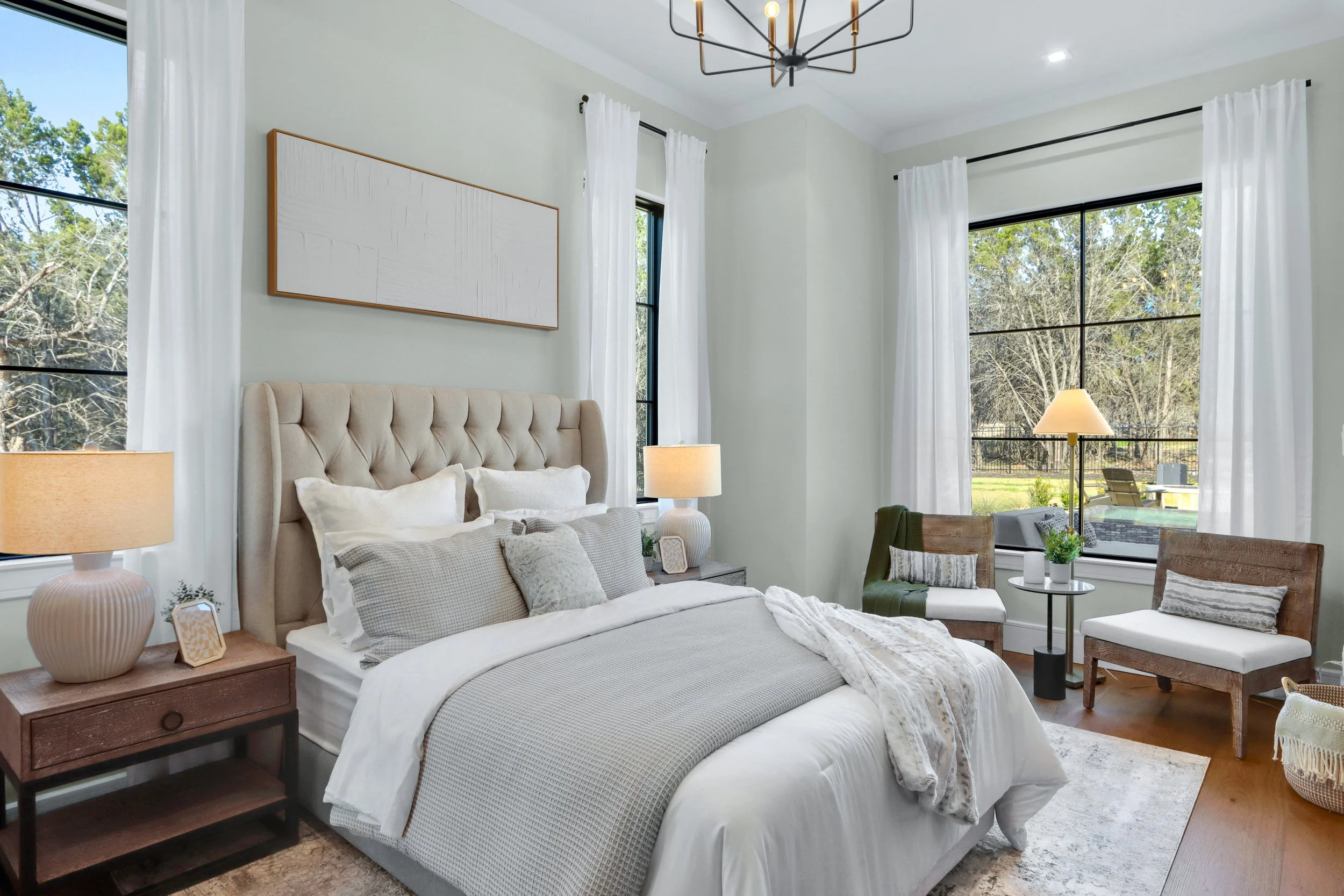 A cozy bedroom with a large bed, beige tufted headboard, white bedding, and gray pillows. Two nightstands with lamps flank the bed, and there are two wooden chairs with cushions near the window. The room has large windows with white curtains, allowin