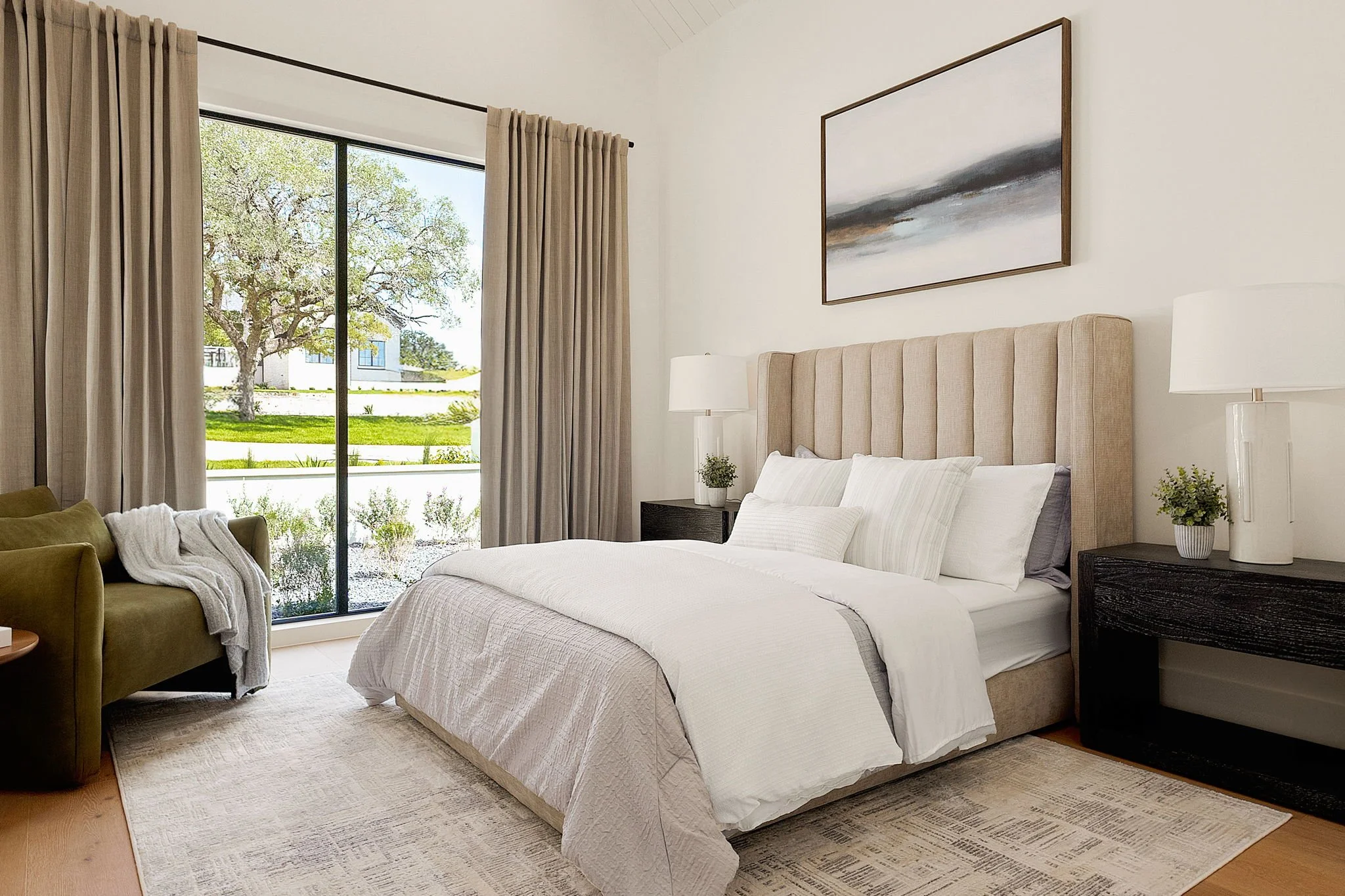 A bedroom with a large bed, beige upholstered headboard, white bedding, and multiple pillows. There are black nightstands with white lamps on each side of the bed, and framed abstract art hangs above it. A sliding glass door with beige curtains leads