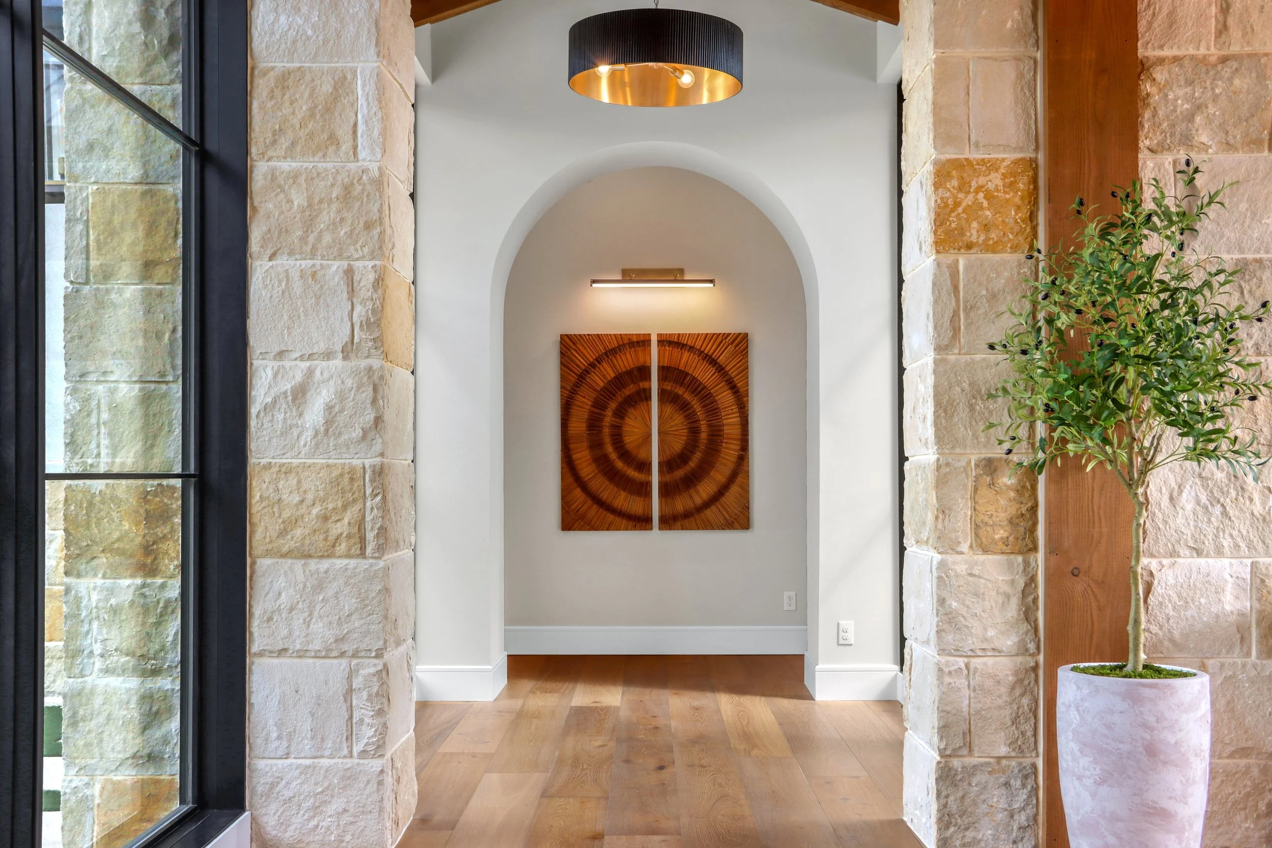 Interior view of a modern home hallway with stone walls, a potted plant, and two paintings of concentric circles on the wall, illuminated by lighting.