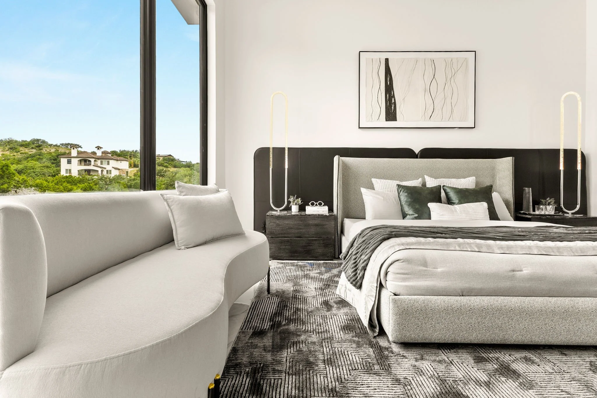 Modern bedroom with large window showing greenery and houses outside, beige upholstered bed with white and dark pillows, black headboard, matching dark bedside table with decorative lamp and accessories, gray and beige abstract art on white wall, gra