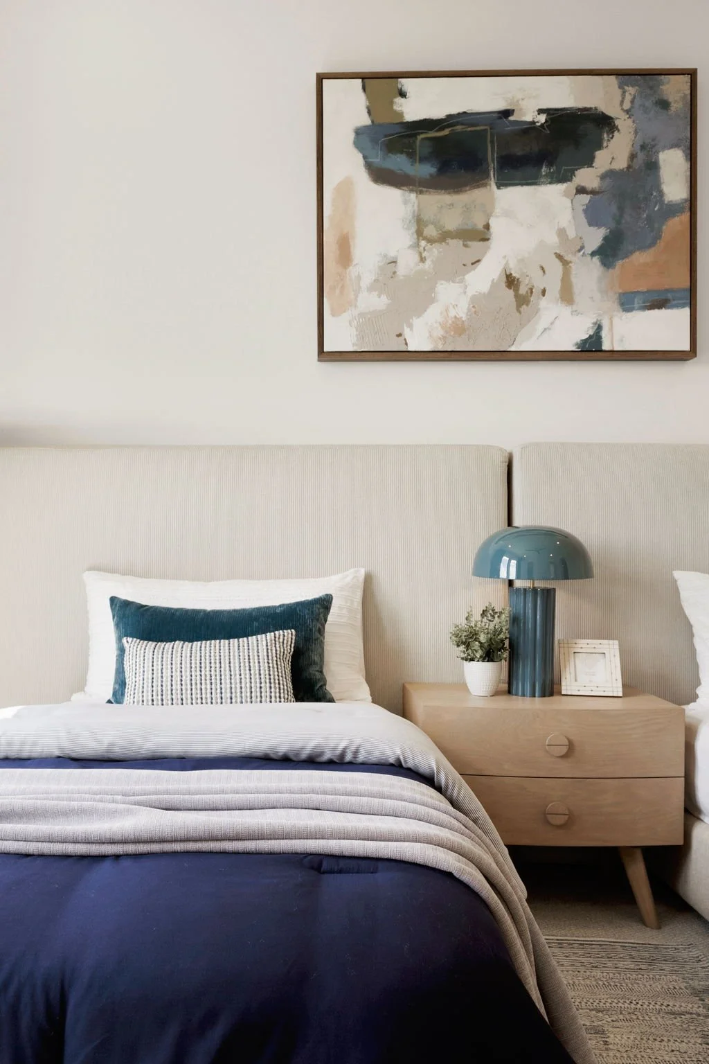 A modern bedroom with a bed, pillows, a nightstand with a blue lamp, a small plant, and a picture frame, with an abstract art painting hanging above.