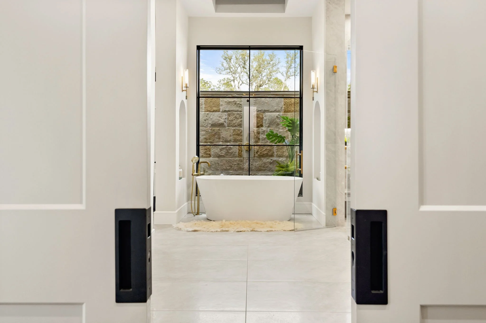 View of a modern bathroom with a freestanding bathtub, large window, stone wall, and indoor plants, seen through partially open white doors.