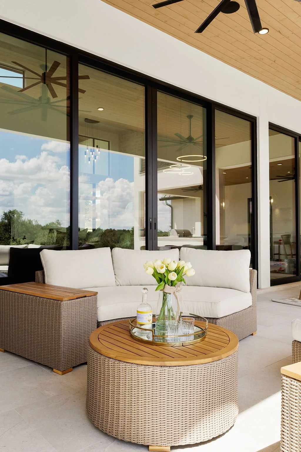Modern outdoor patio with wicker furniture, a white sofa, a round wicker table with a wooden top, a vase of white tulips, and glassware, adjacent to a glass-walled house with visible ceiling fans and decorations.