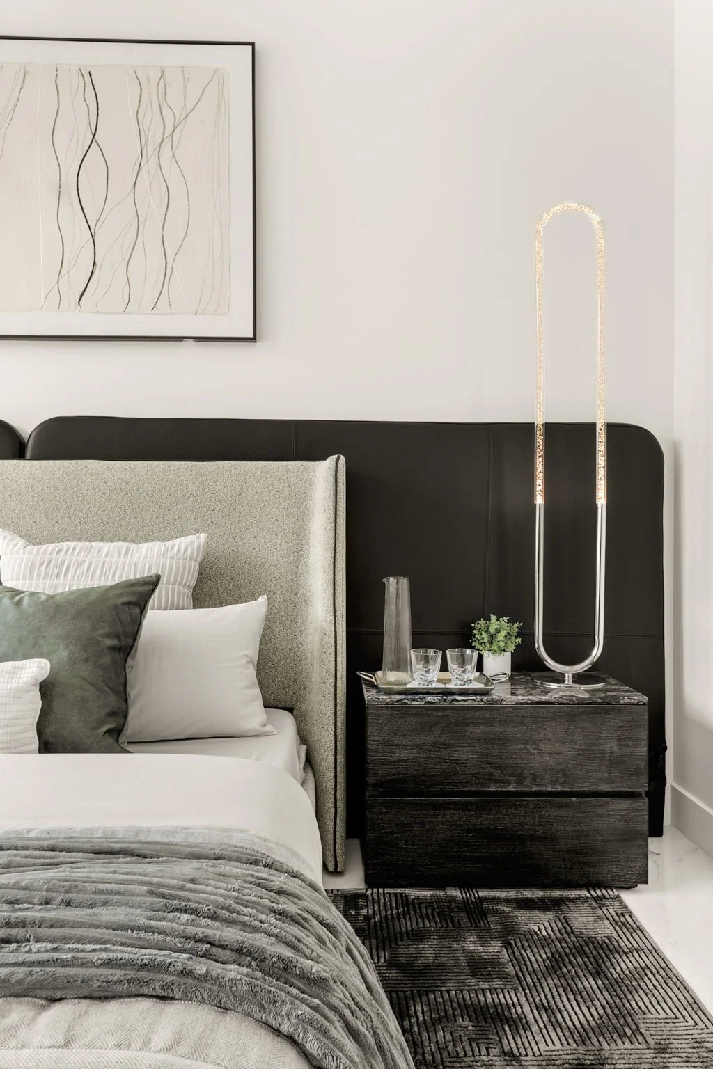Modern bedroom with a bed featuring white and gray pillows, a textured beige headboard, a dark wooden nightstand with a glass pitcher, glasses, and a small plant, a black padded wall panel, and an artistic framed line drawing on the white wall. A tal